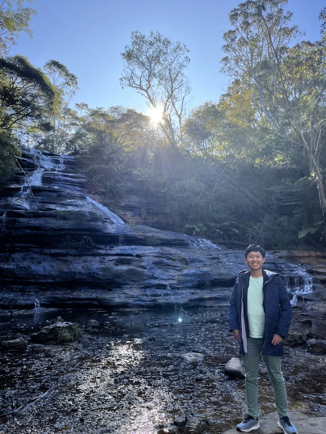 Standing at the base of a cascading waterfall in the Blue Mountains with sun streaming through trees