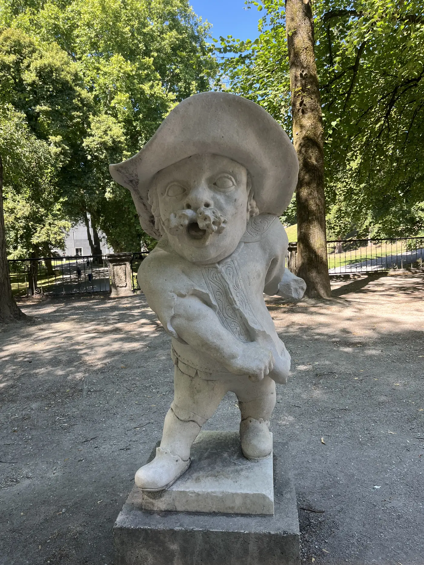Weathered stone dwarf statue with a wide hat in the Dwarf Garden of Mirabell Gardens, Salzburg, surrounded by tall trees