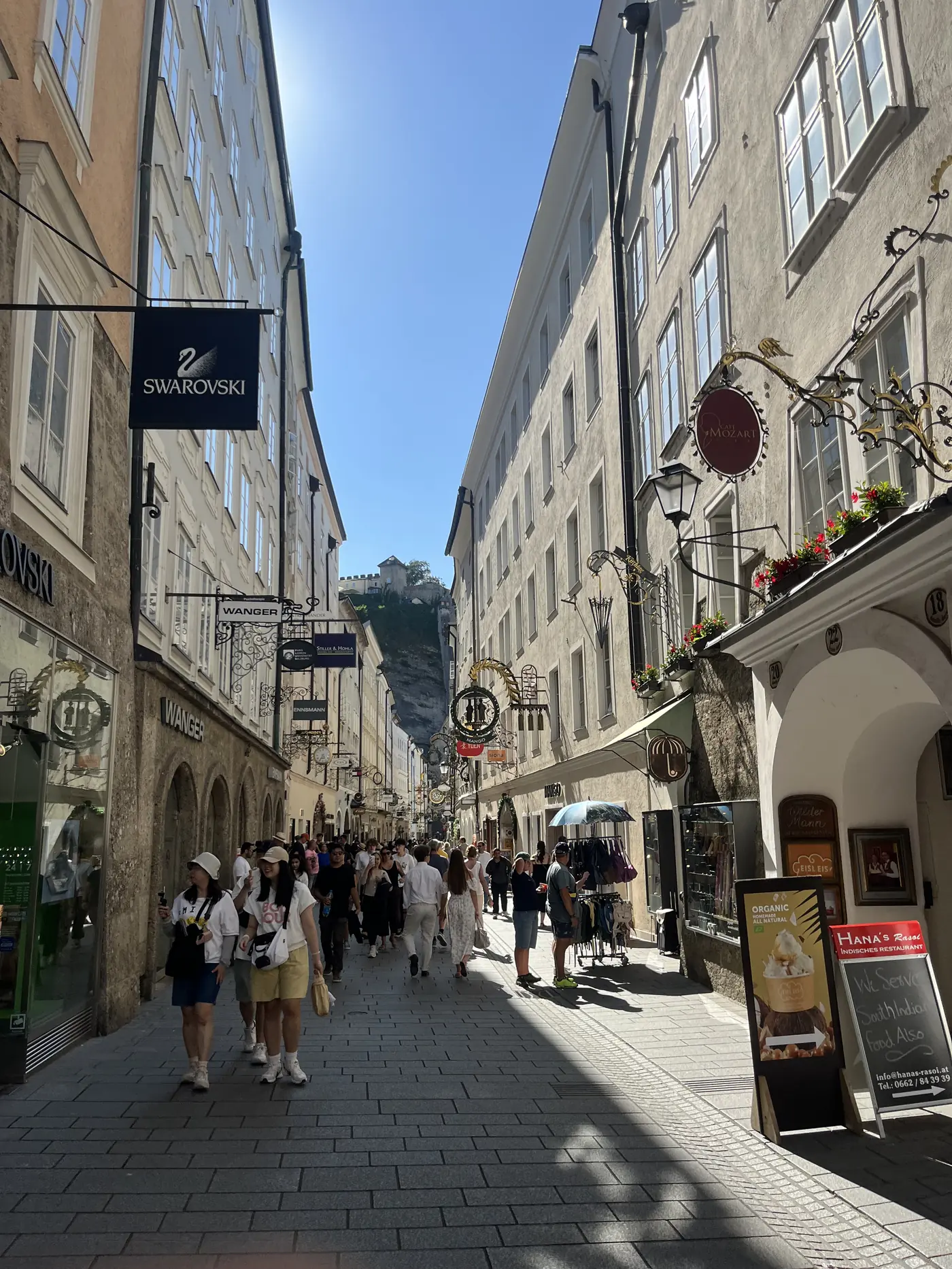 Getreidegasse shopping street in Salzburg with ornate wrought-iron guild signs, a Swarovski storefront, and Hohensalzburg Fortress visible atop the cliff at the end