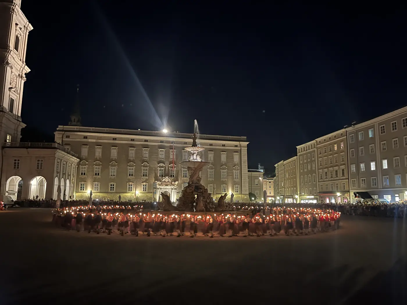 Nighttime torch-lit procession circling the Residenzbrunnen fountain in Salzburg's Residenzplatz, performers in red traditional dress