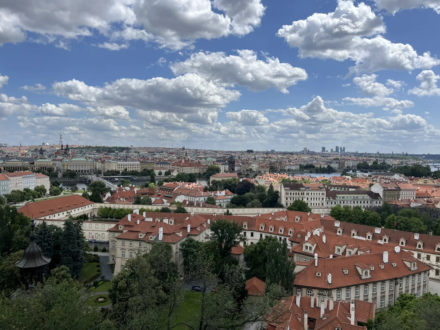 Panoramic view over Prague's red-roofed rooftops toward the Vltava river, bridges, and the city skyline under a cloudy sky
