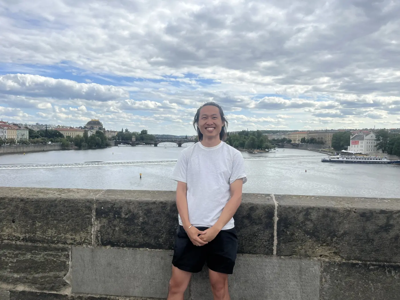 Shin standing on Charles Bridge with the Vltava river, bridges, and Prague skyline behind him under a cloudy sky