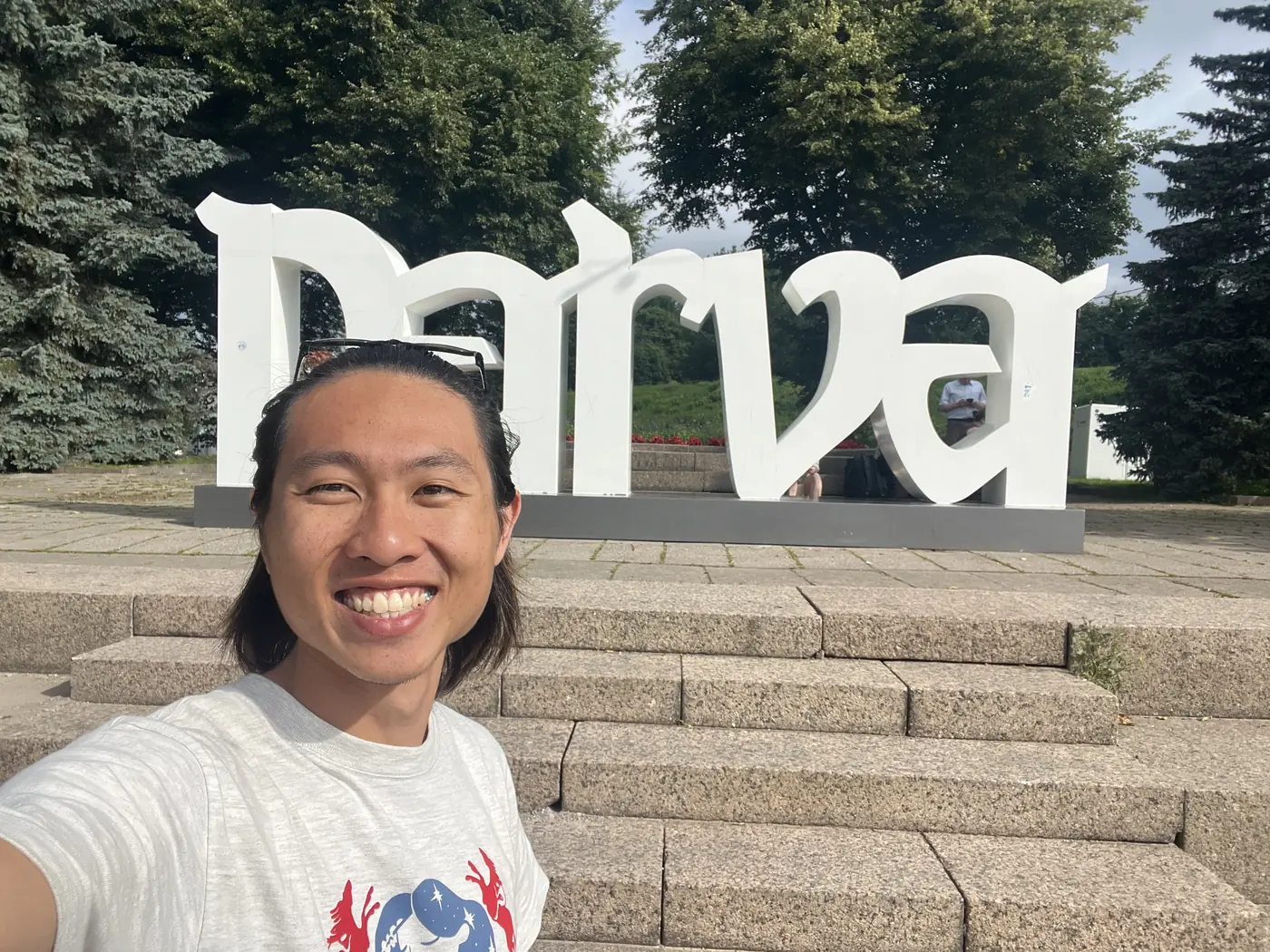 Shin selfie in front of a giant white NARVA sign on stone steps, with green trees behind, at the Estonian border city of Narva