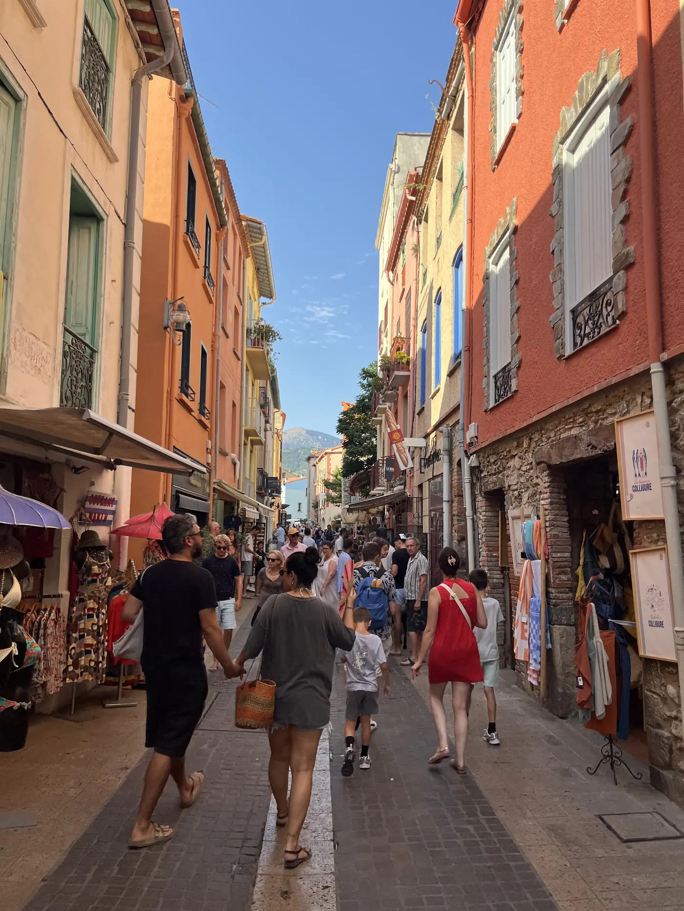 Colourful narrow street in Collioure lined with pink and orange buildings, shops, and morning crowds