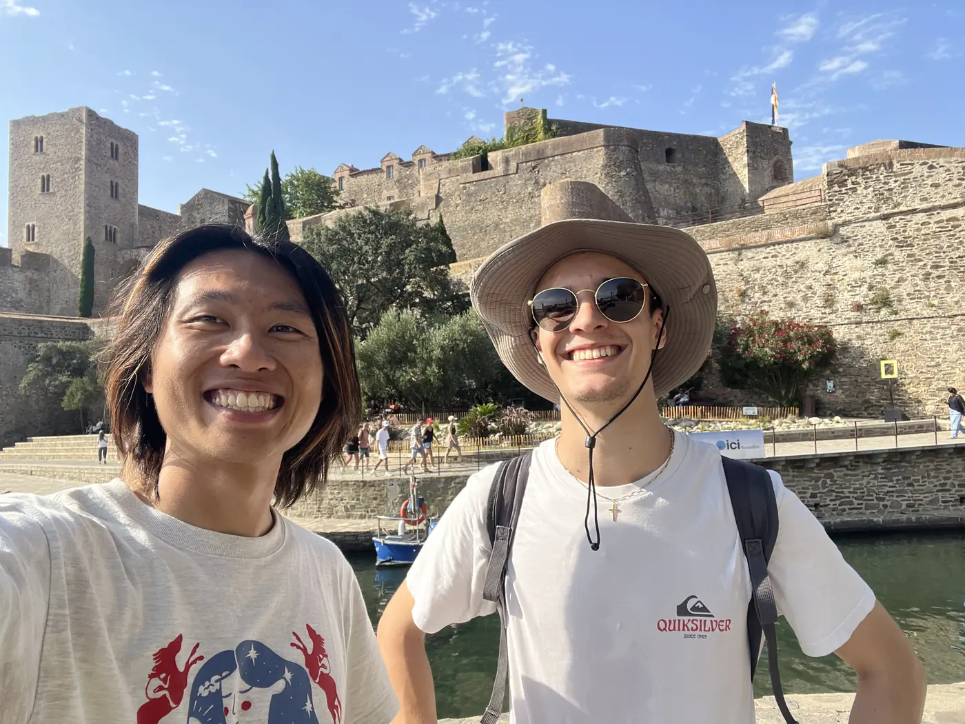 Shin and a friend in a sunhat smiling in front of the Château Royal de Collioure, harbour and boats visible behind them