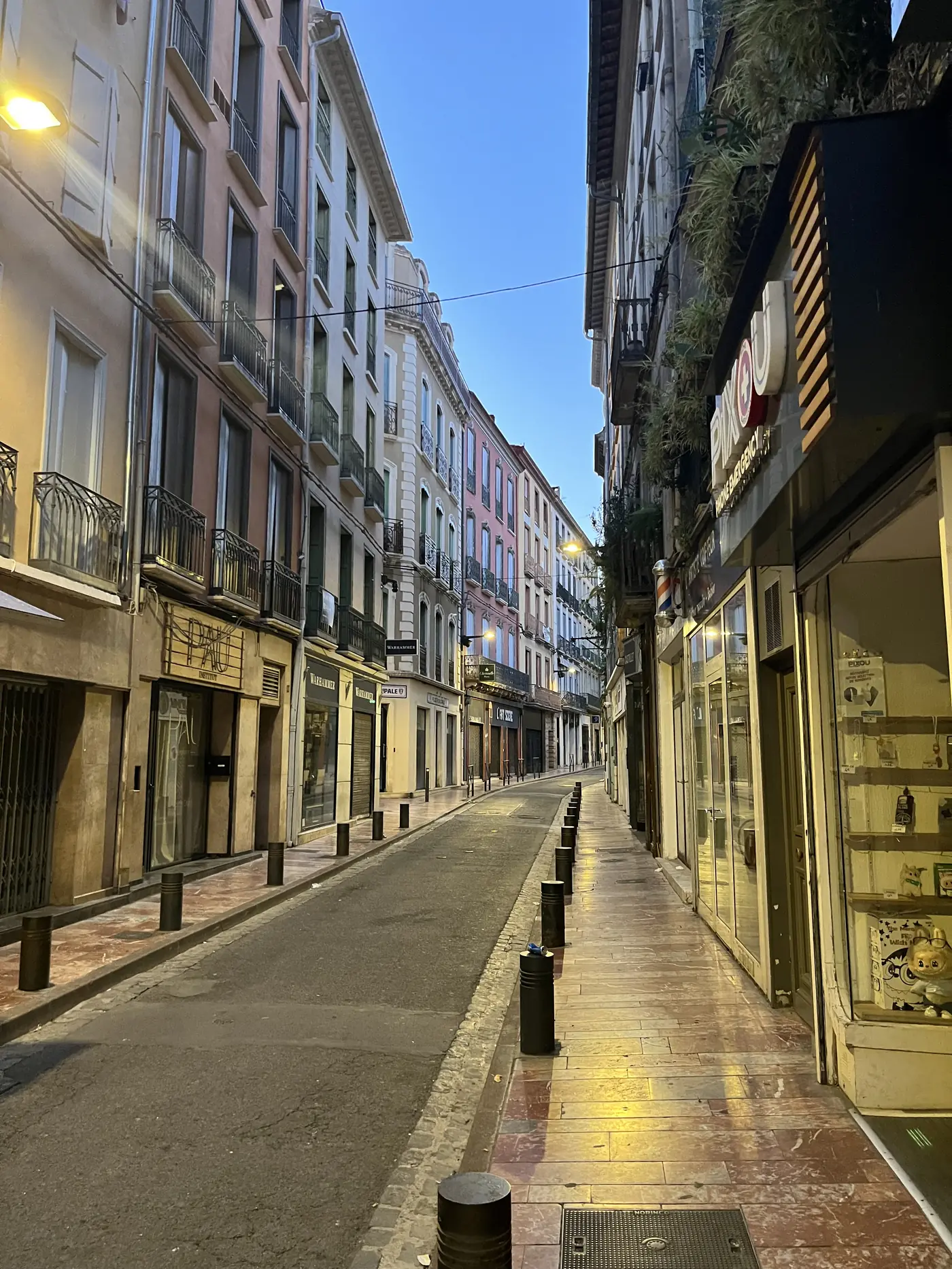 Empty Perpignan old town street at sunrise, pastel-coloured buildings and shop awnings glowing in golden morning light