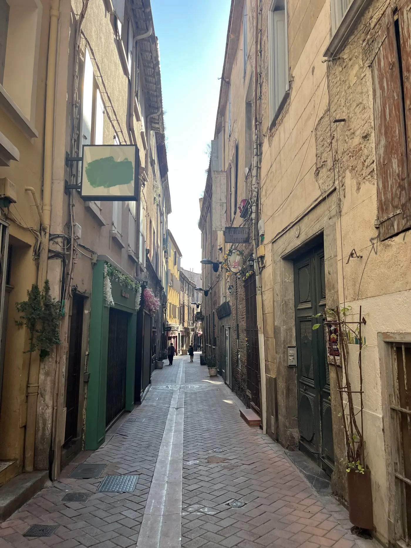 Narrow medieval alley in Perpignan's old town, warm stone buildings and green shop shutters, empty in the midday sun