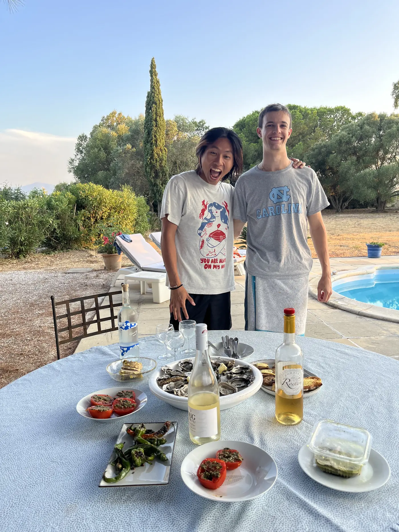 Shin and a friend smiling by a poolside table set with oysters, tomatoes, olives, and white wine at sunset near Perpignan