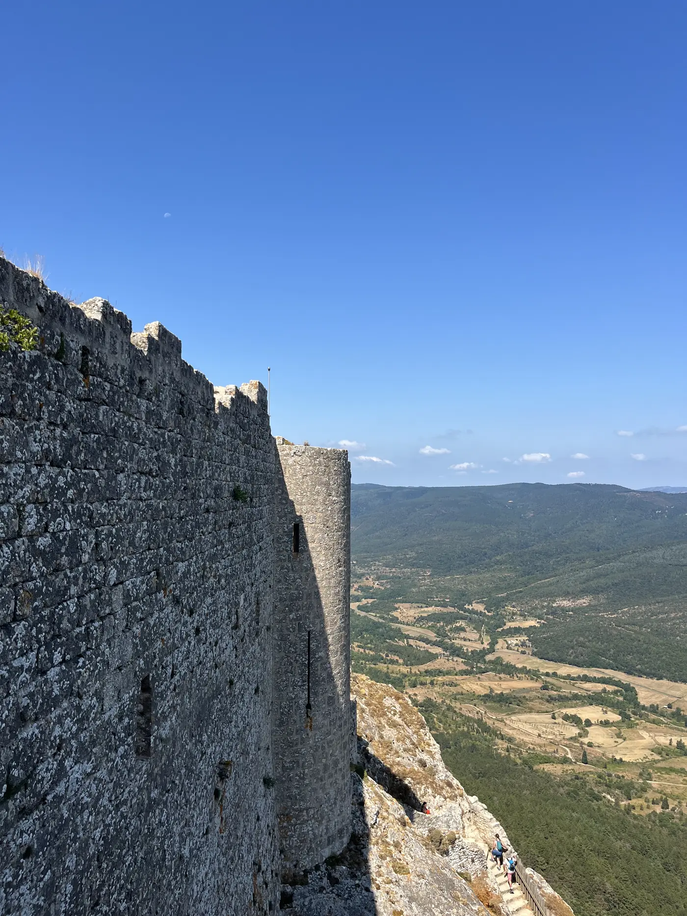 Ancient stone battlements of Château de Peyrepertuse running along a cliff edge, with a vast green valley far below