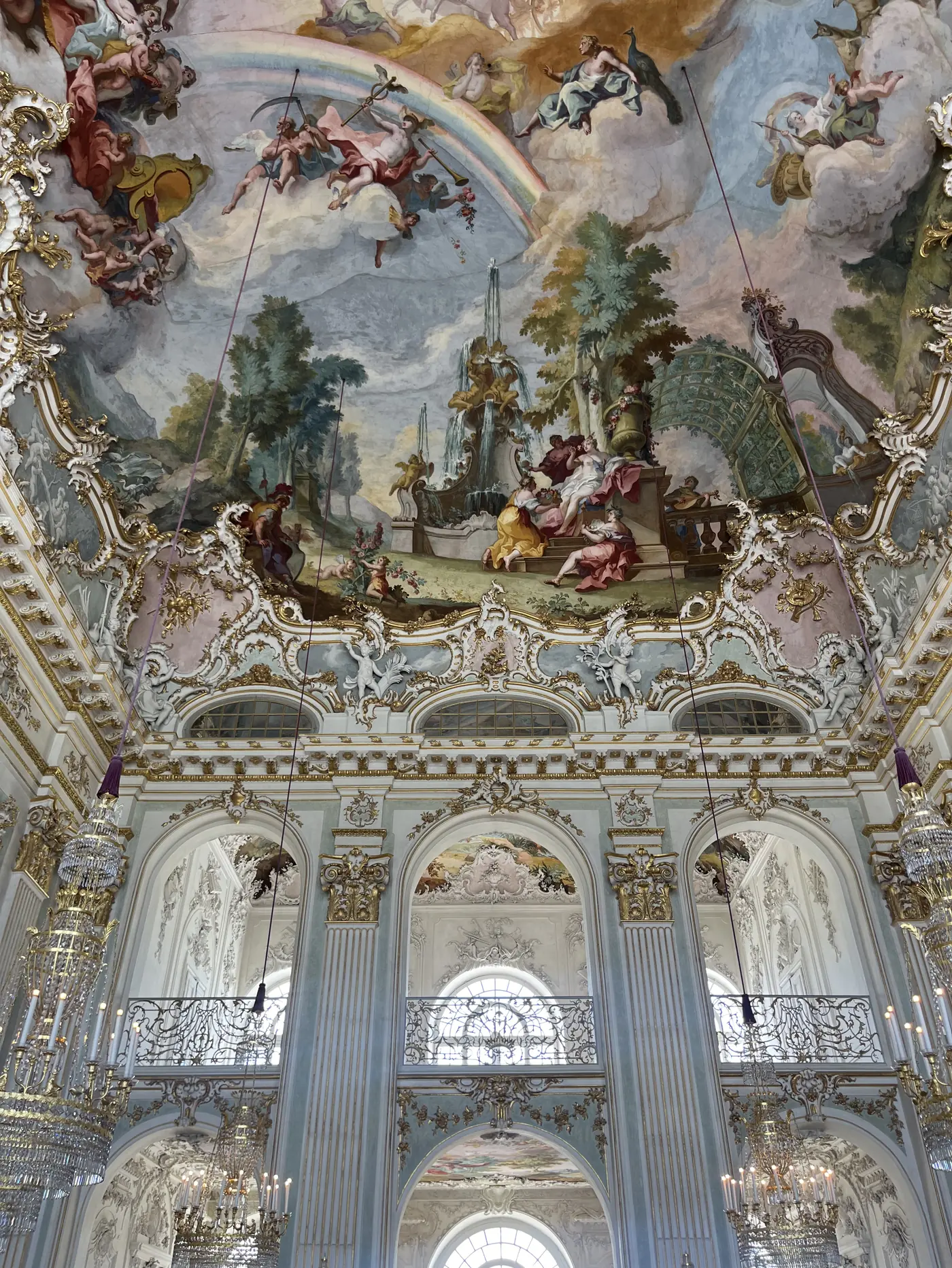 Ornate rococo ceiling fresco inside a Munich palace hall with painted gods and cherubs, gold stucco, and crystal chandeliers below