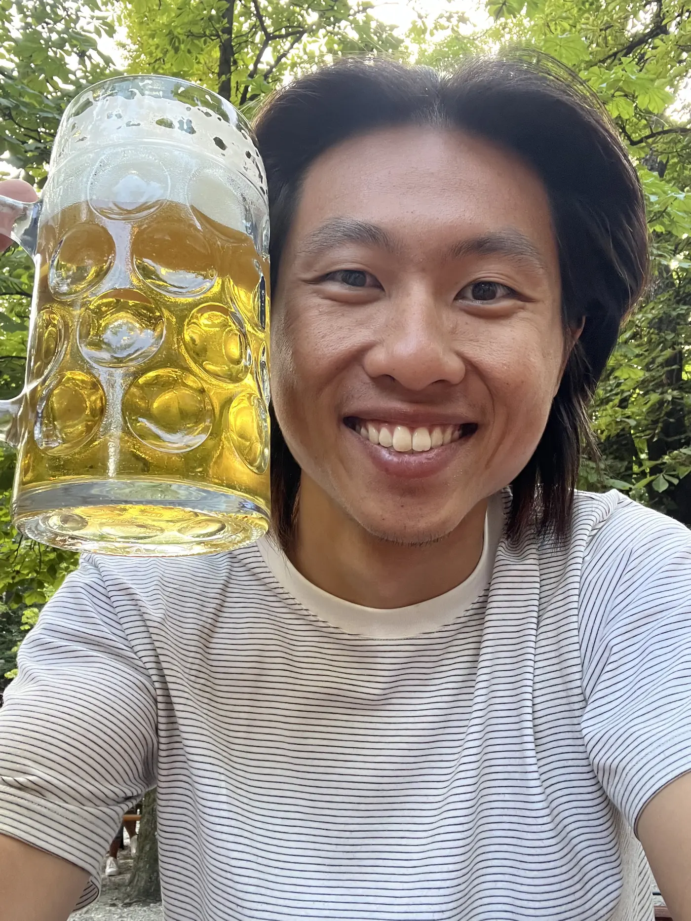 Shin selfie holding up a full one-liter Maß beer stein under the green canopy of a beer garden