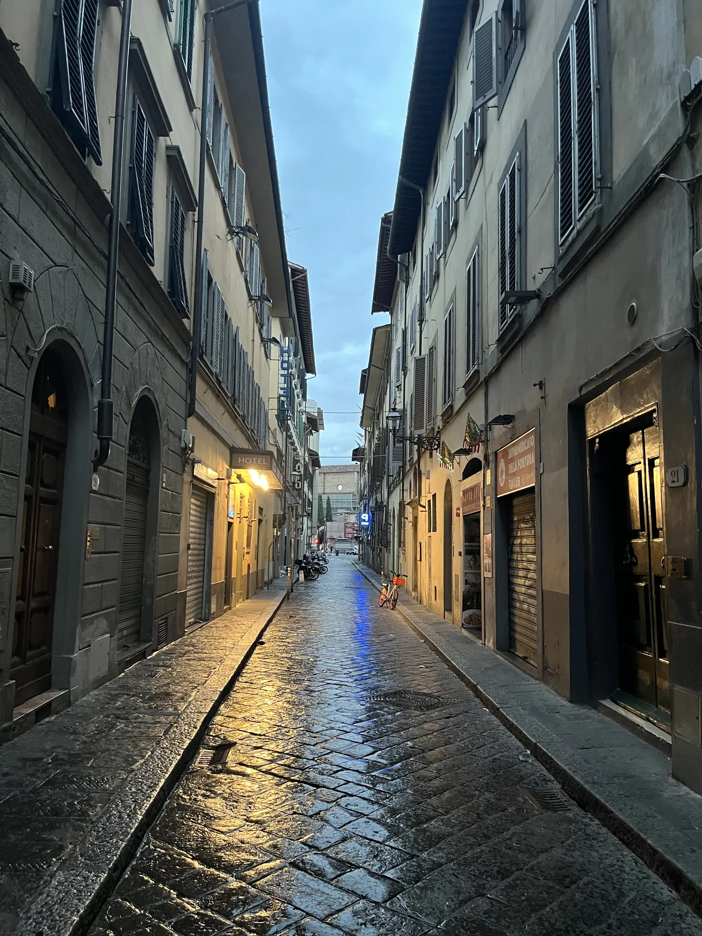 Empty narrow Florence street glistening with rain at dusk, warm shop lights reflecting off the wet cobblestones