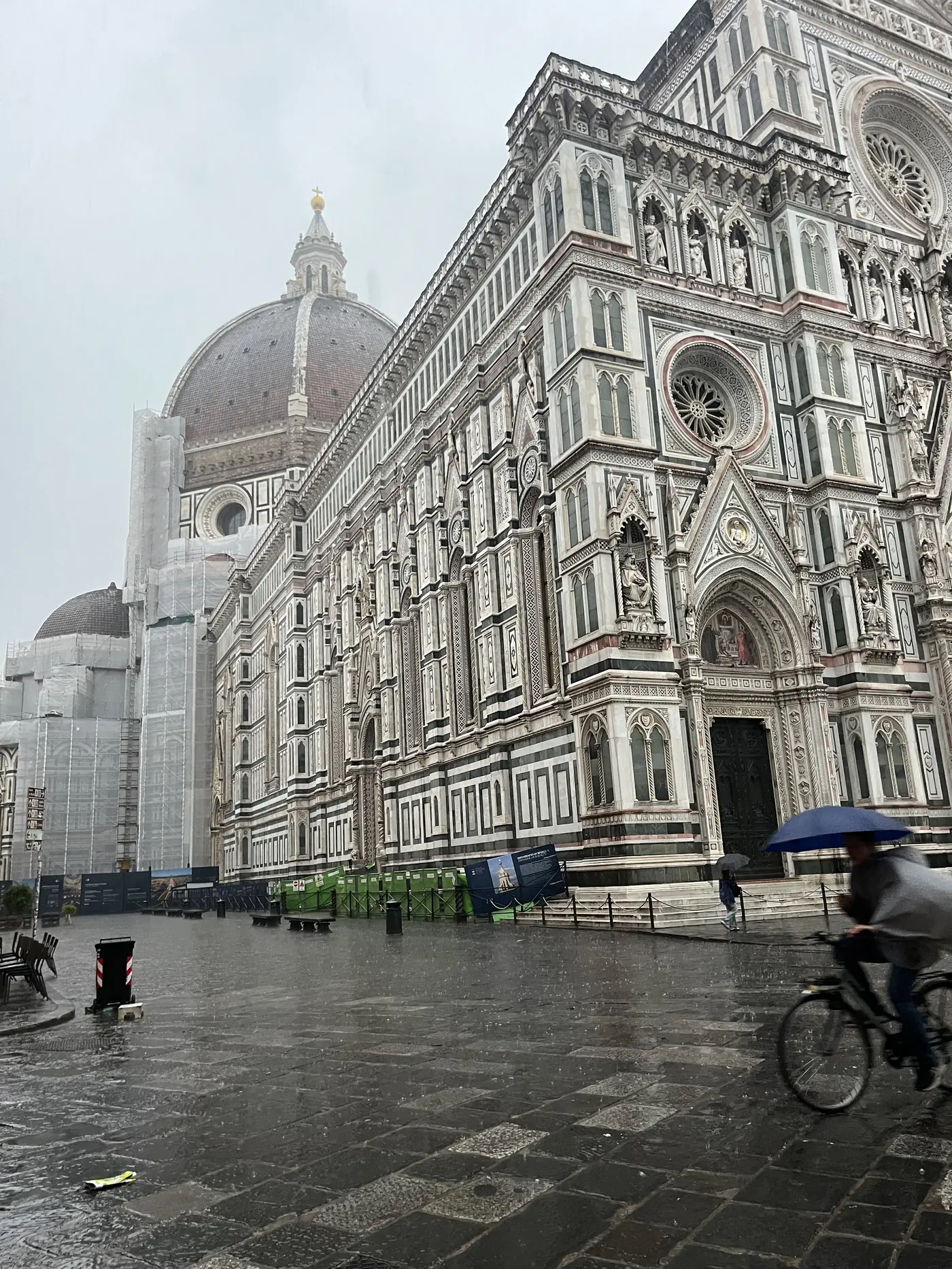 Florence Cathedral (Il Duomo) with its striped marble facade and dome in the rain, a cyclist passing with an umbrella