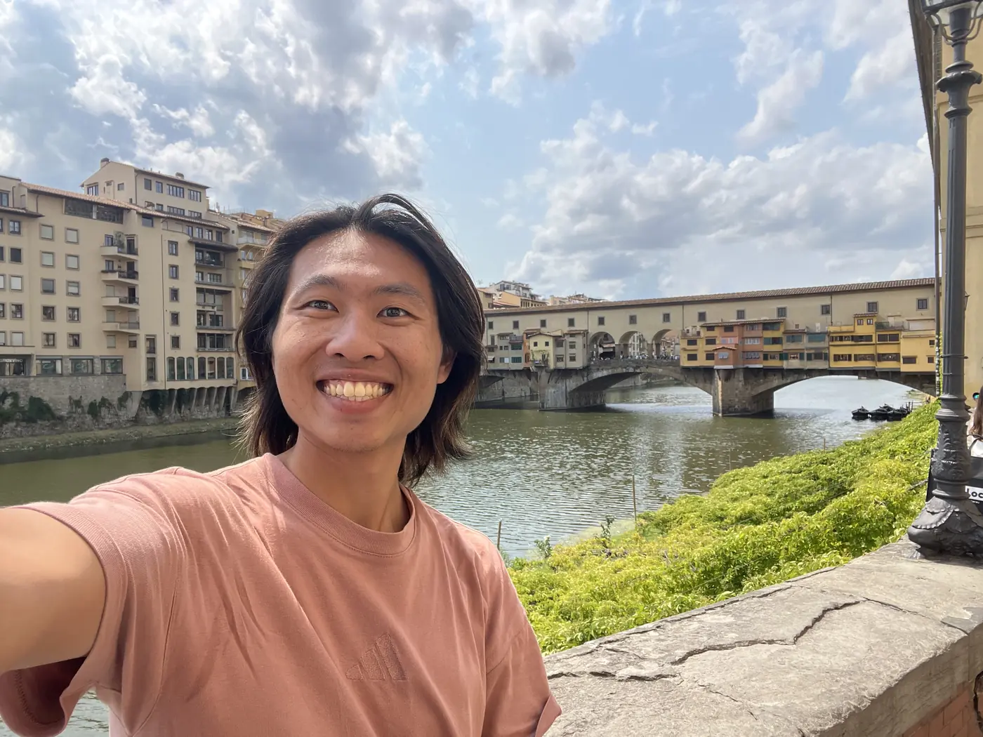 Shin selfie by the Arno river with the Ponte Vecchio's shop-lined bridge visible behind him under a partly cloudy sky