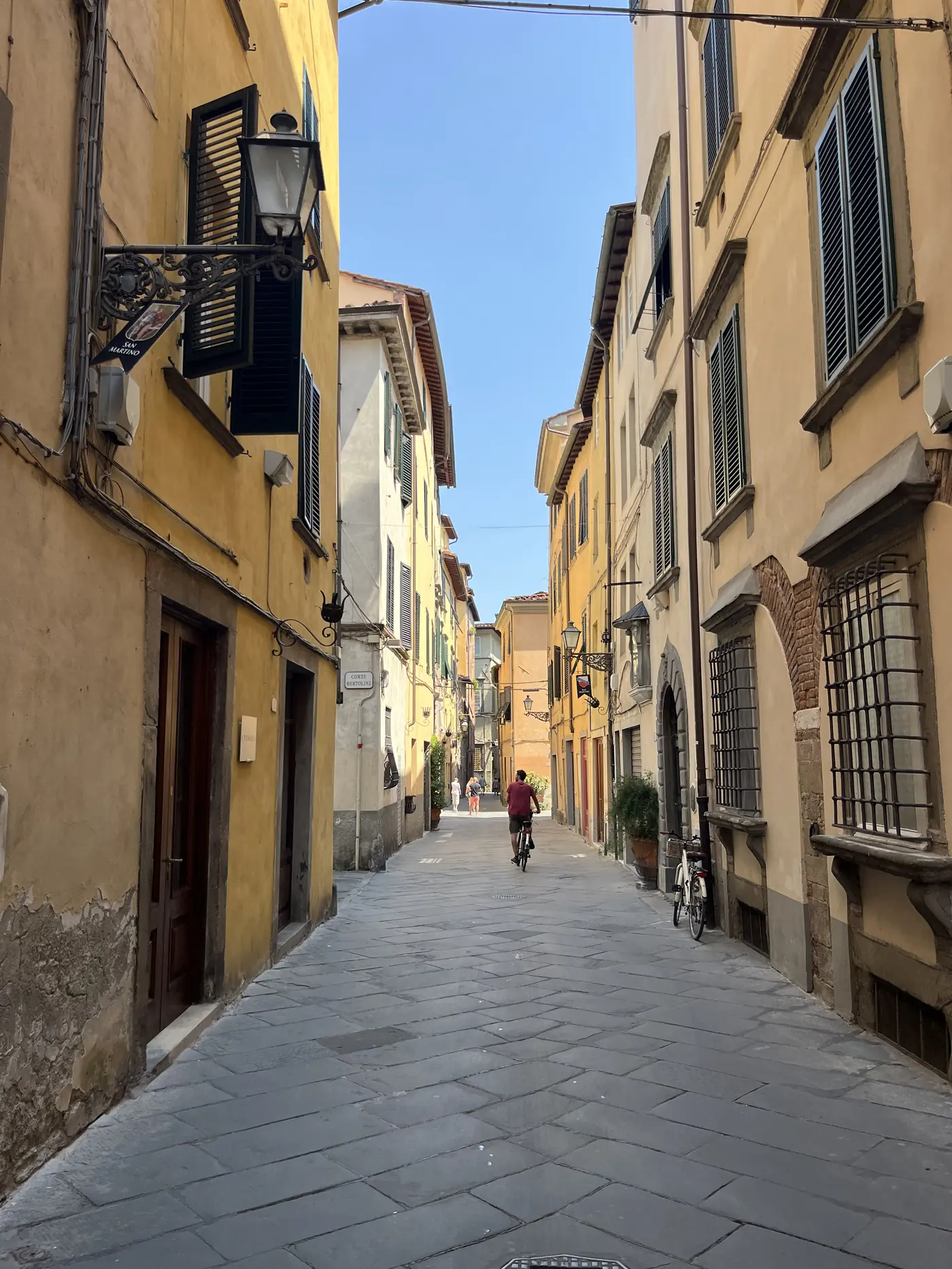 Narrow warm-toned street inside Lucca's old town with a cyclist riding past shuttered windows and hanging lanterns