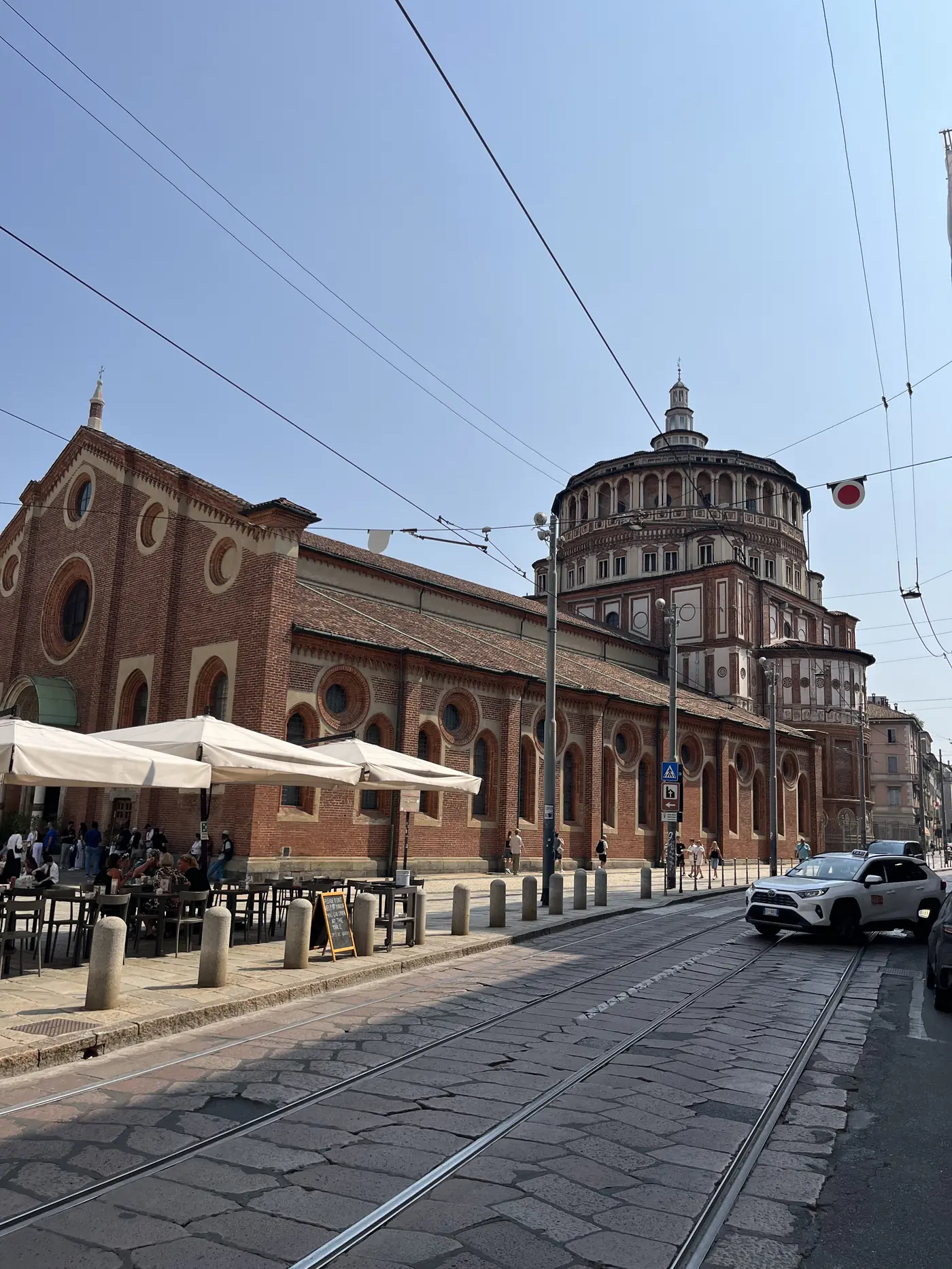 Street view of Santa Maria delle Grazie in Milan, the red-brick basilica's nave and domed apse, with a café terrace and tram tracks in front