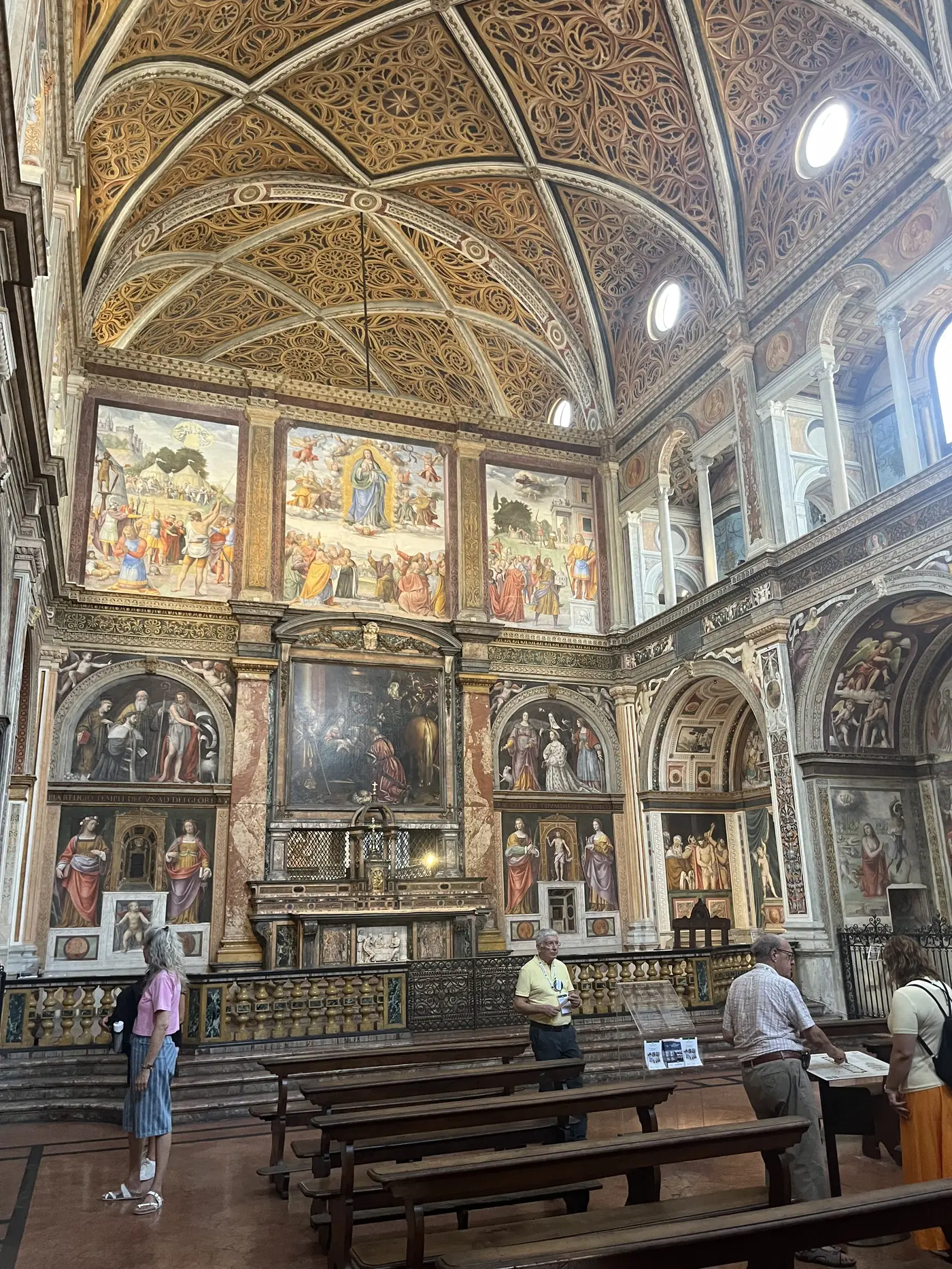 Ornate Renaissance chapel interior in Milan with a gilded coffered ceiling, frescoed walls depicting religious scenes, and rows of wooden pews