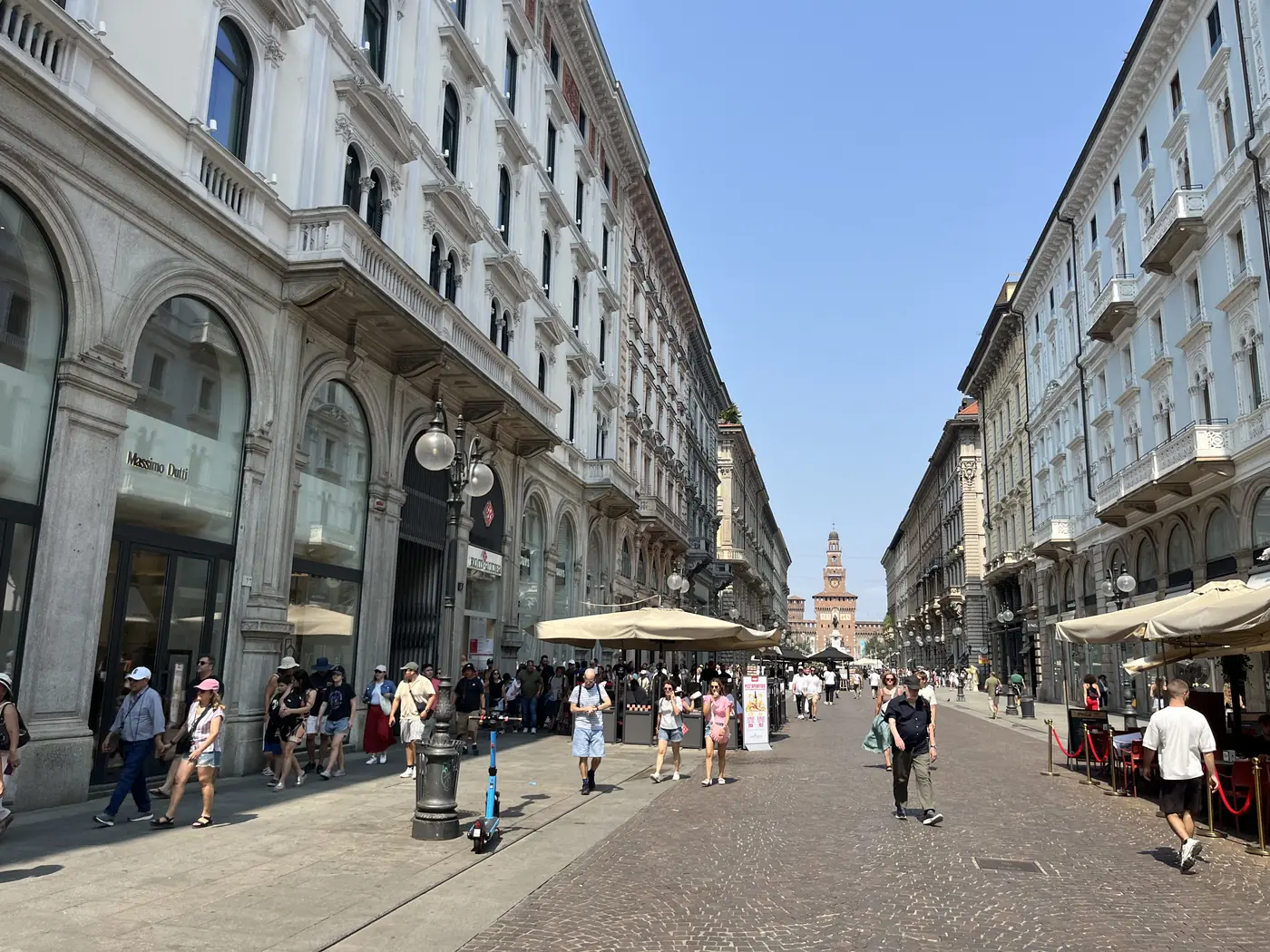 Elegant pedestrian shopping street in central Milan lined with arcaded 19th-century buildings, with a castle tower visible in the distance