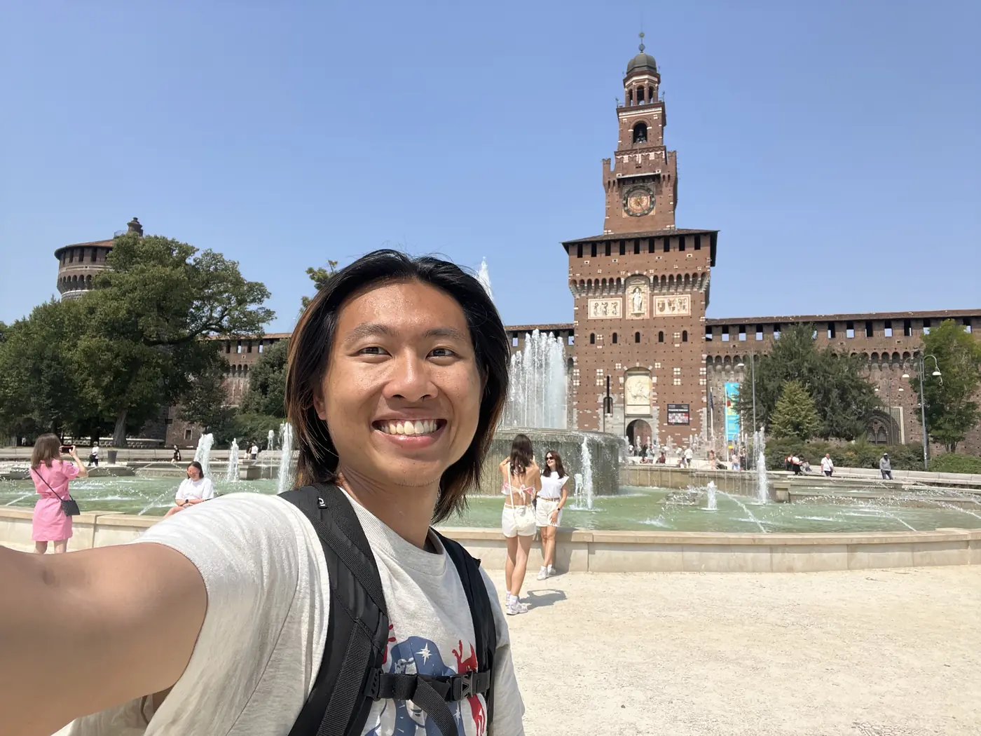 Shin selfie in front of the Sforza Castle's brick fortress towers and a large fountain in Piazza Castello, Milan