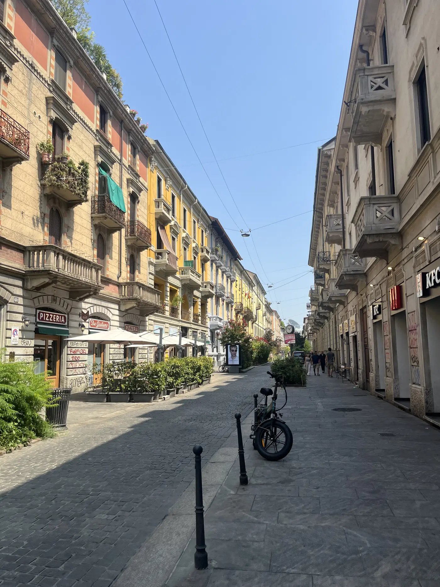 Quiet Milan street lined with ornate 19th-century apartment buildings, wrought-iron balconies, a corner pizzeria, and a parked cargo bike