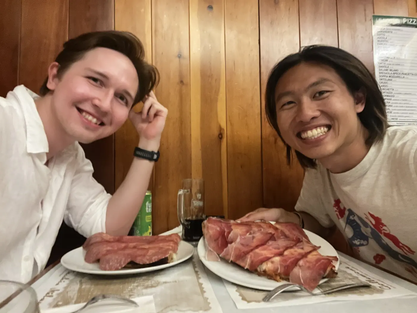 Shin and a friend grinning at a restaurant table with two plates of sliced grilled meat and prosciutto