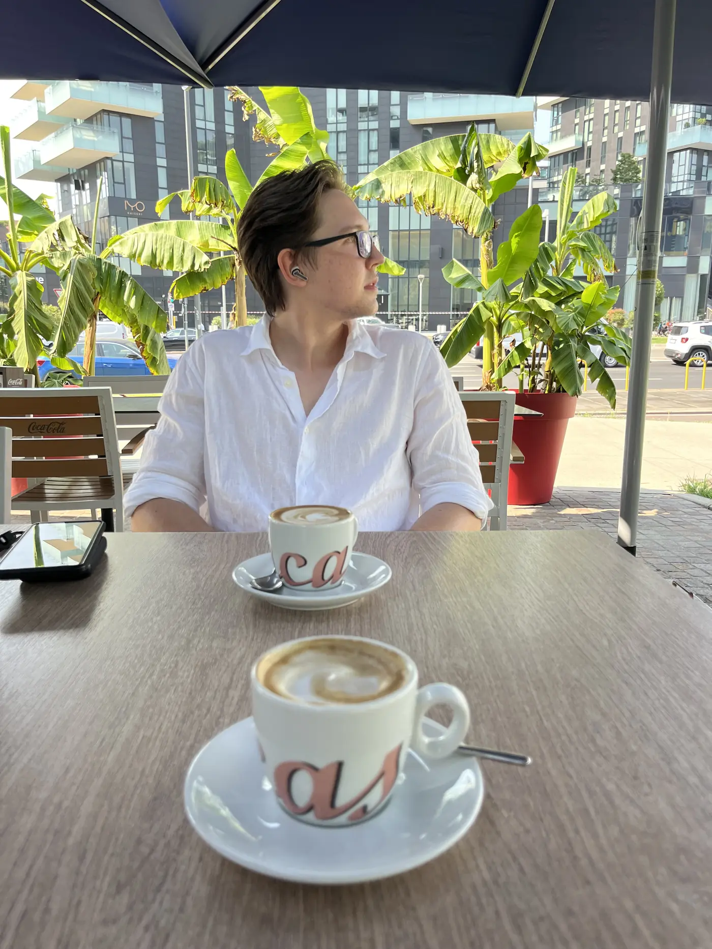 A friend sitting at an outdoor café table in Milan with two cappuccinos, banana palm trees, and modern glass apartment buildings behind