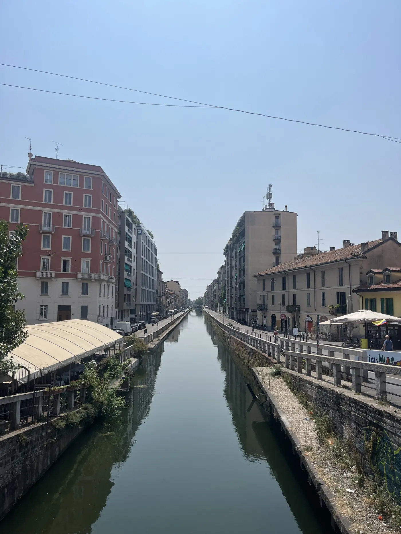 The Naviglio Grande canal in Milan's Navigli district, lined with apartment buildings, cafes, and a covered terrace, calm reflective water