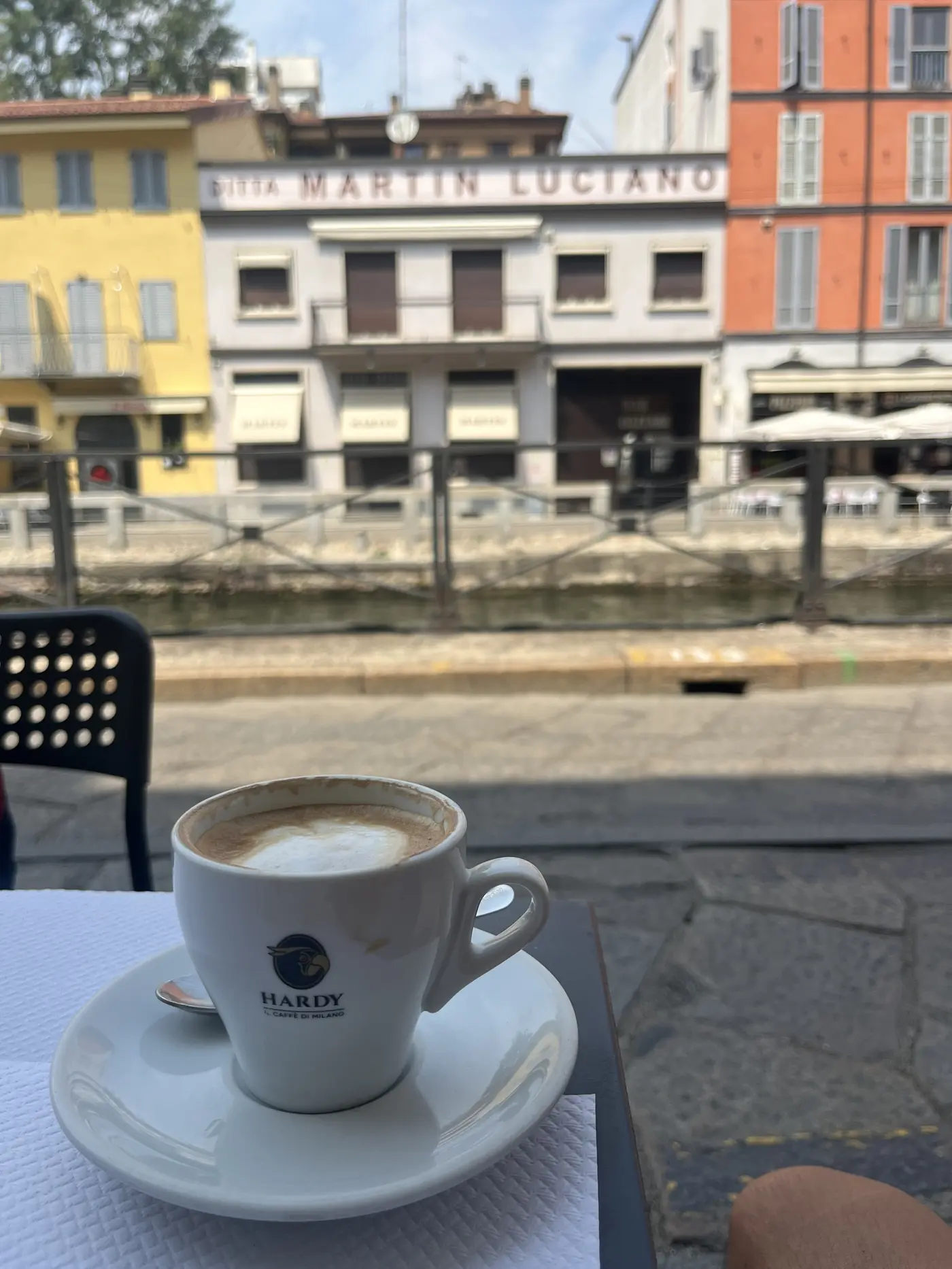 A cappuccino on a café table along the Navigli canal in Milan, colourful buildings and the canal railing visible behind