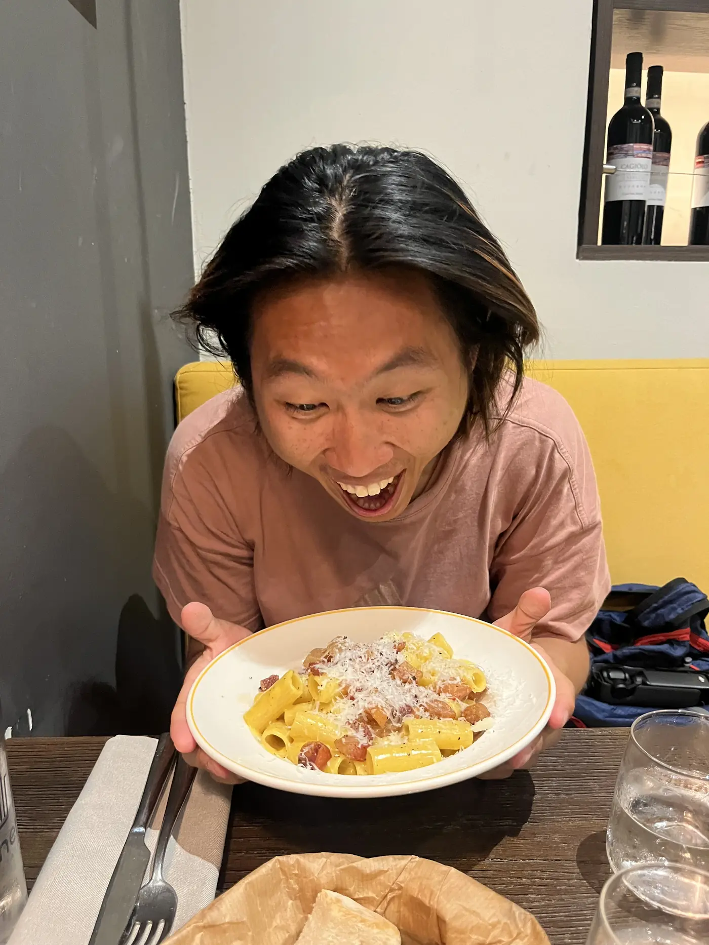 Shin grinning wide-eyed while holding a plate of rigatoni carbonara-style pasta at a Milan restaurant table