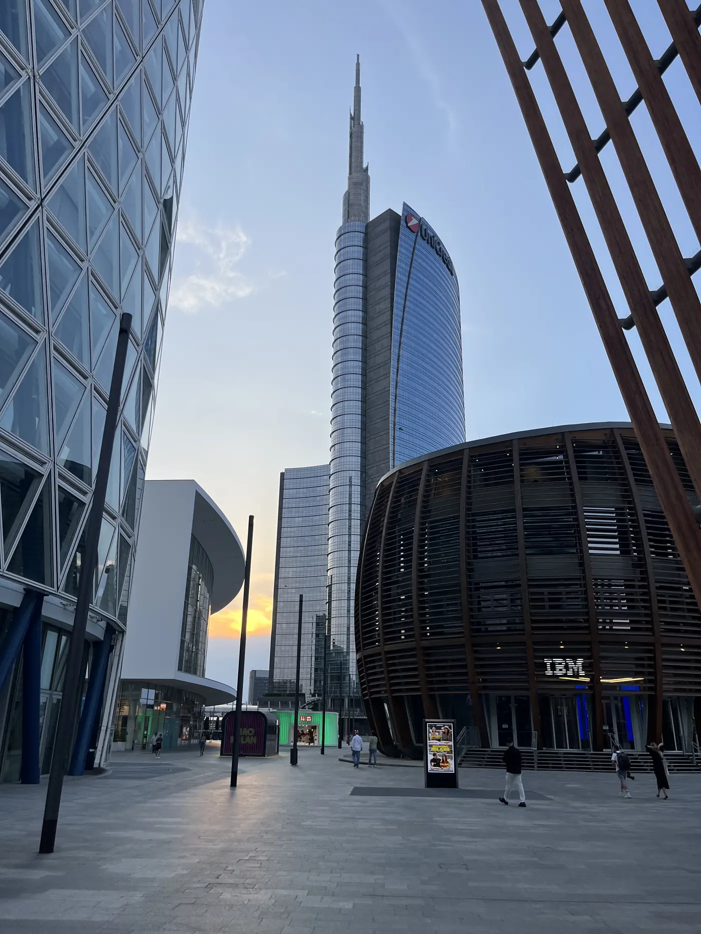 Milan's Porta Nuova skyline at dusk, the UniCredit Tower's tapering spire silhouetted against a sunset glow between glass buildings