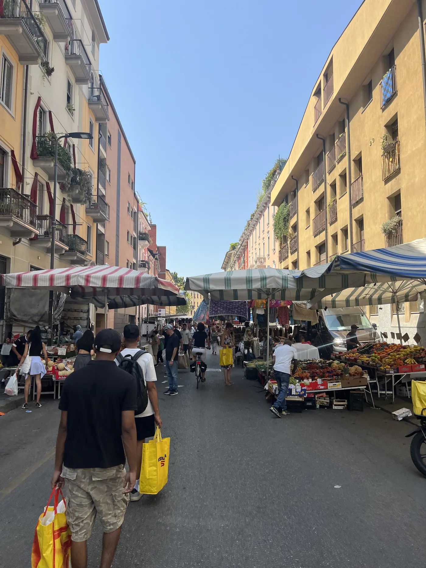 A busy neighbourhood street market in Milan with striped awnings, fruit and vegetable stalls, and shoppers carrying bags