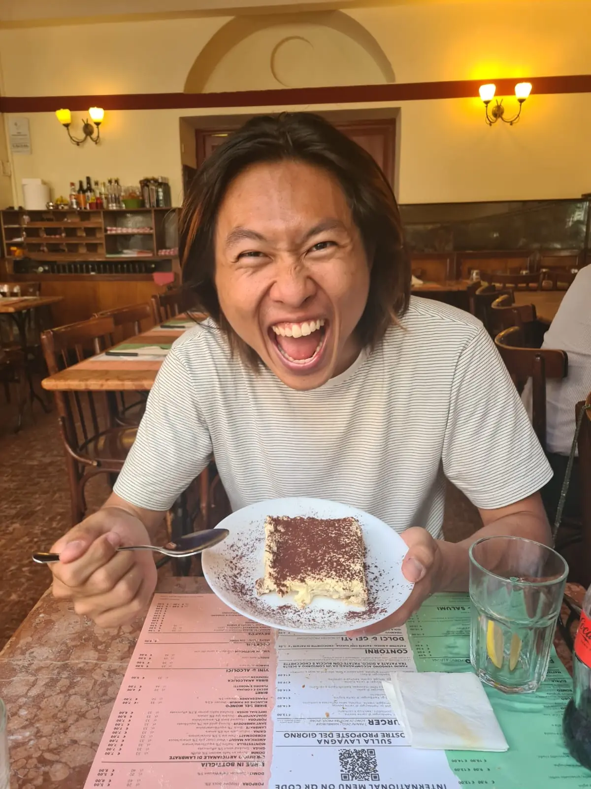 Shin laughing with his mouth wide open, holding a plate of tiramisu and a spoon at a classic Italian trattoria