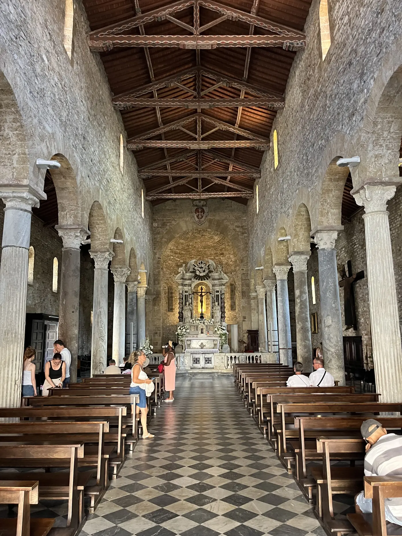 Romanesque church interior in Pisa's Piazza dei Miracoli with rows of stone columns, a wooden truss ceiling, and worshippers seated in wooden pews