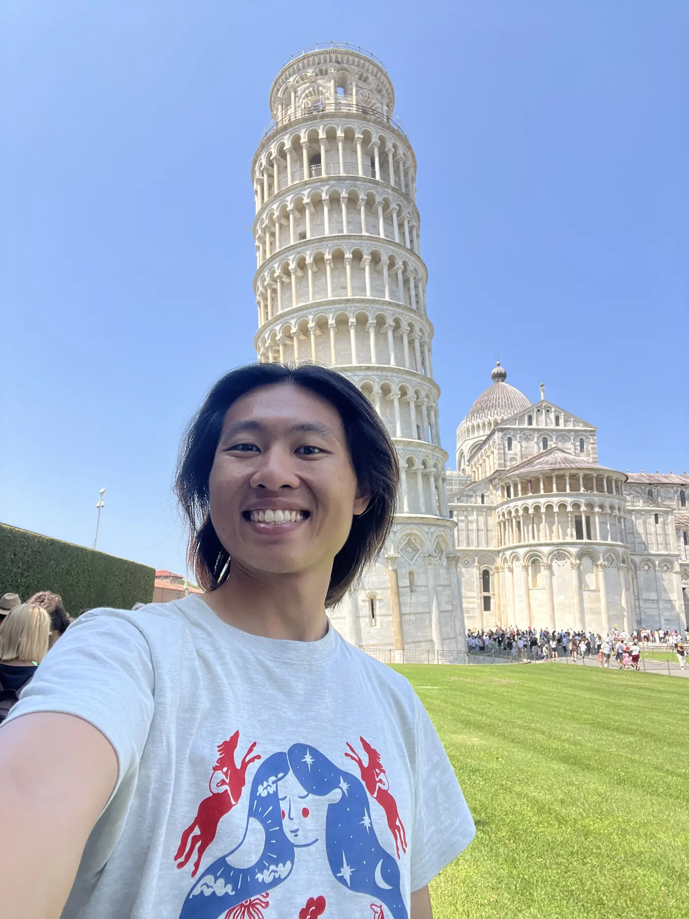 Shin selfie in front of the Leaning Tower of Pisa, the tower appearing to tilt sharply behind him under a clear sky