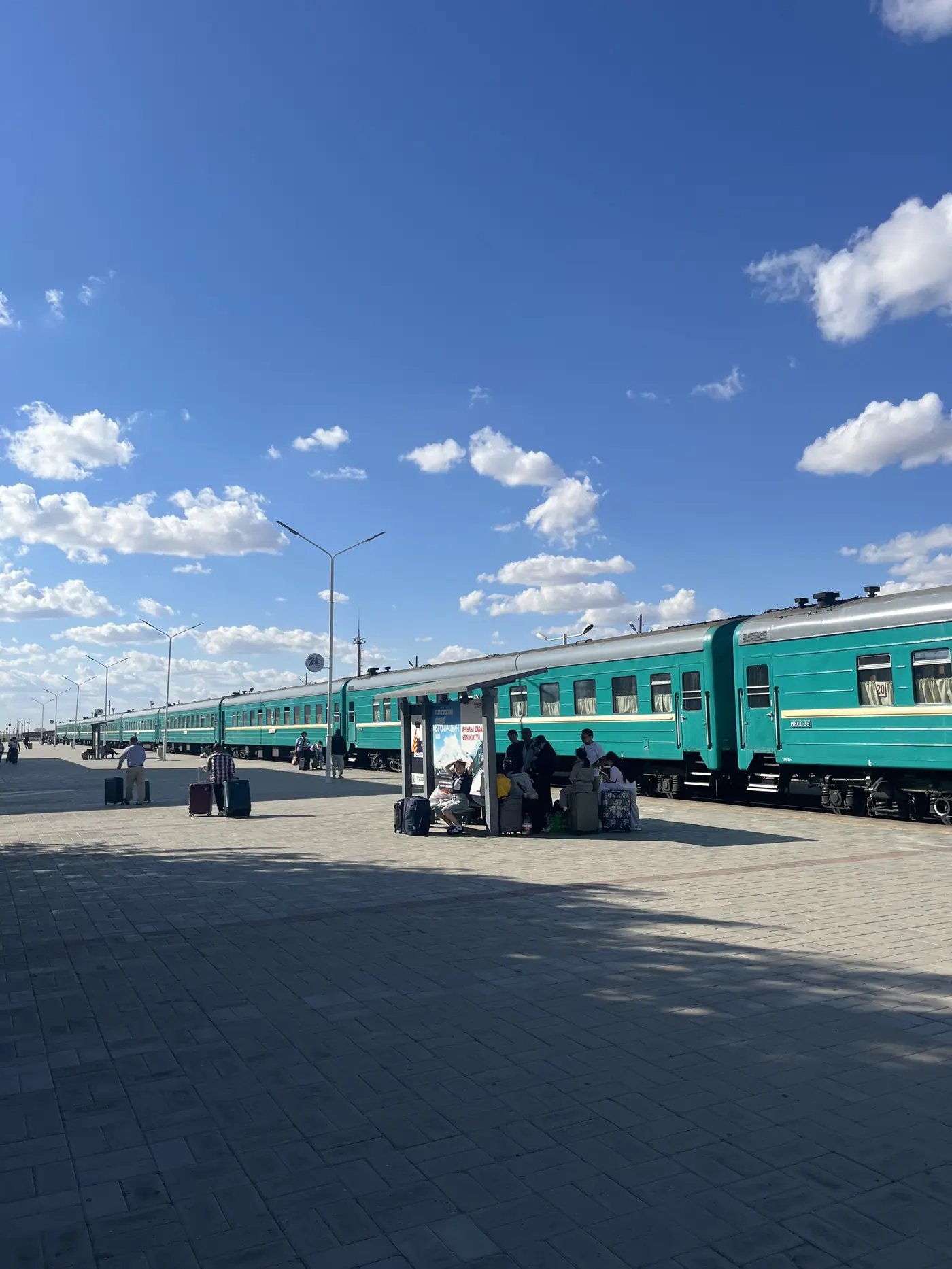 Teal Trans-Mongolian Railway carriages at Zamyn Uud border station platform, passengers with luggage under a vast Mongolian sky