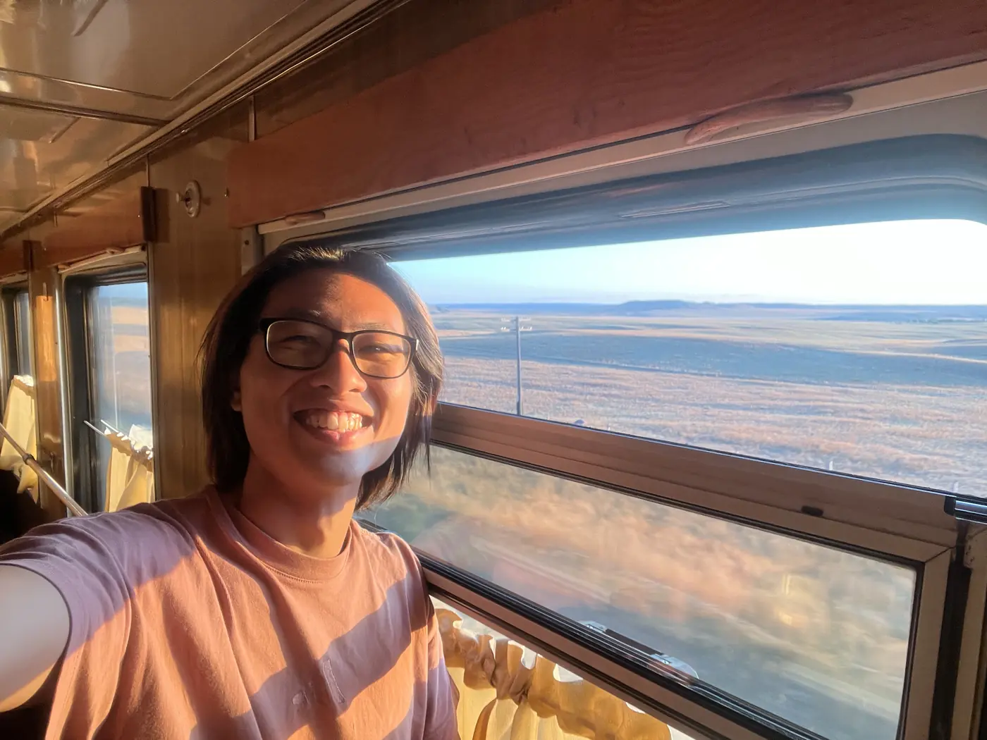 Solo traveller Shin smiling in Trans-Mongolian Railway carriage with Gobi Desert steppe visible through window at golden hour