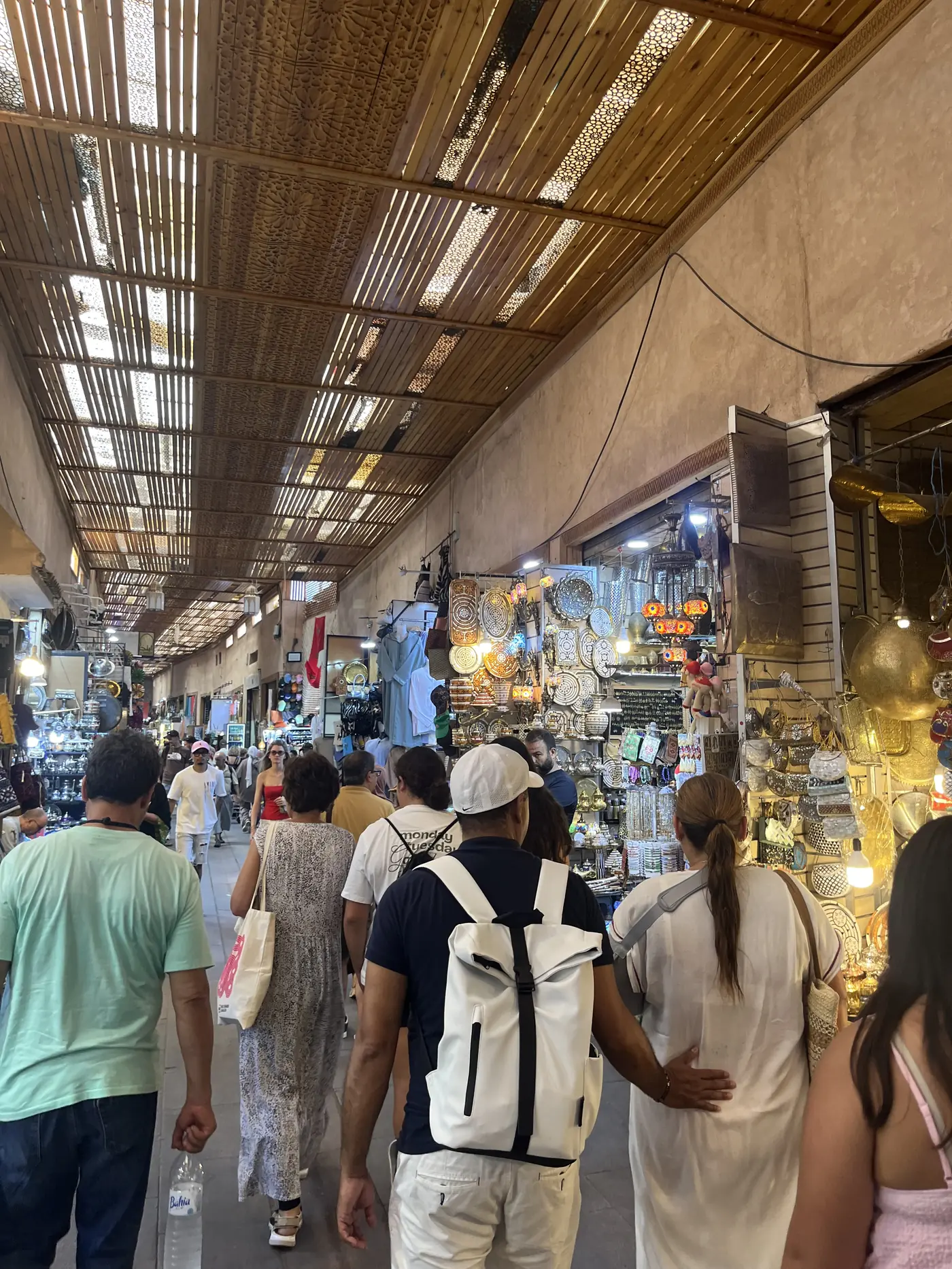 A covered souk alley in Marrakech with a woven reed roof filtering light onto stalls selling lanterns, rugs, and metalwork