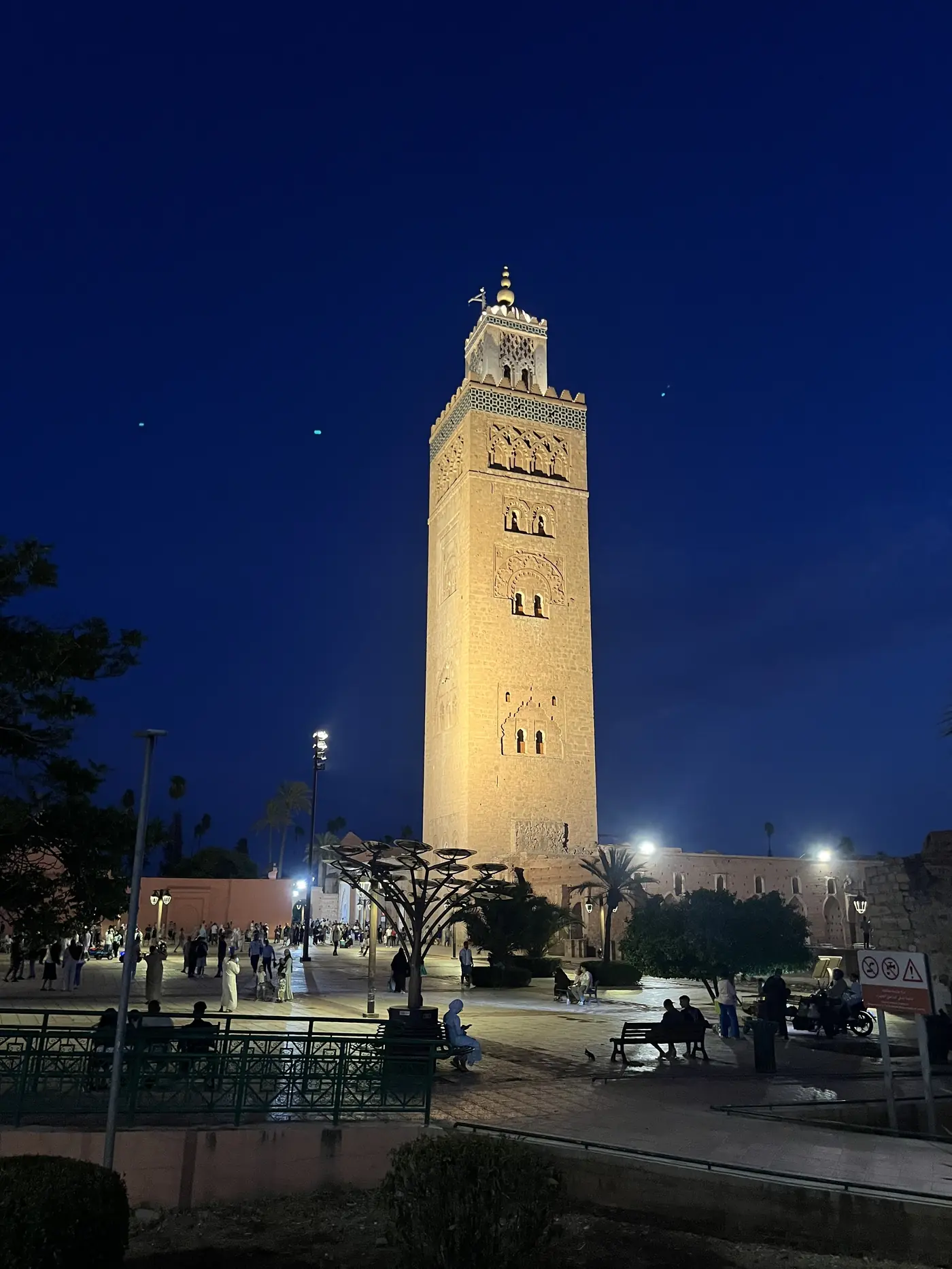 The Koutoubia Mosque's minaret illuminated at night against a deep blue sky, with people gathered in the gardens below