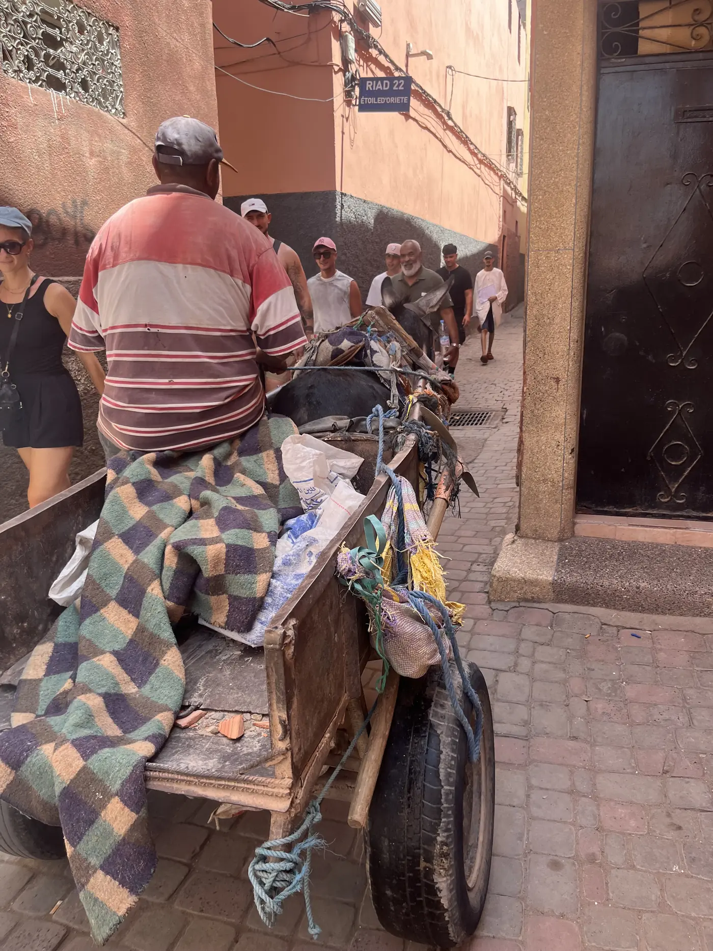 A man pulling a loaded handcart through a narrow Marrakech medina alley past a Riad 22 street sign, tourists following behind