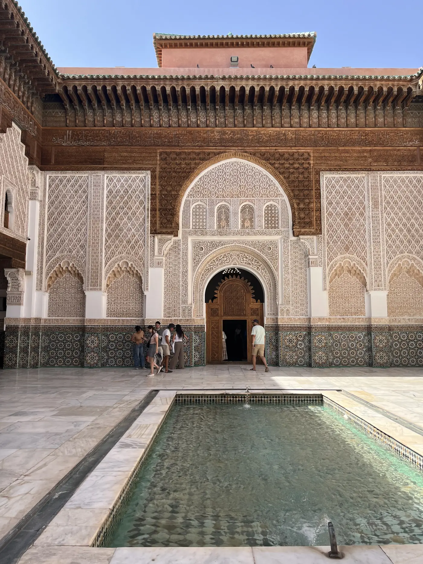 The ornate carved plasterwork and zellige-tiled courtyard of the Ben Youssef Madrasa in Marrakech, with a reflecting pool in the foreground