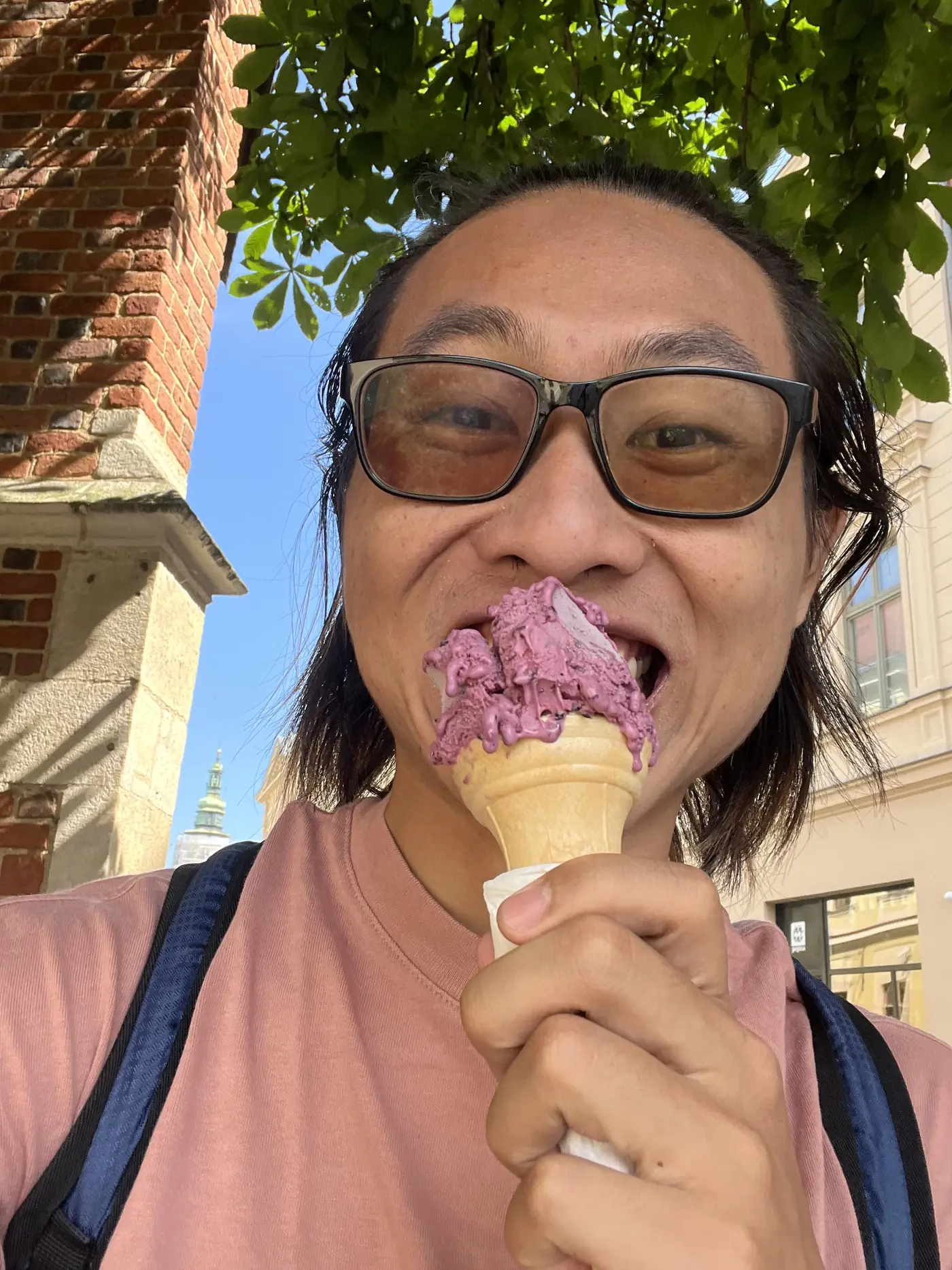 Shin biting into a scoop of purple ice cream on a waffle cone, brick tower and Kraków street visible behind him