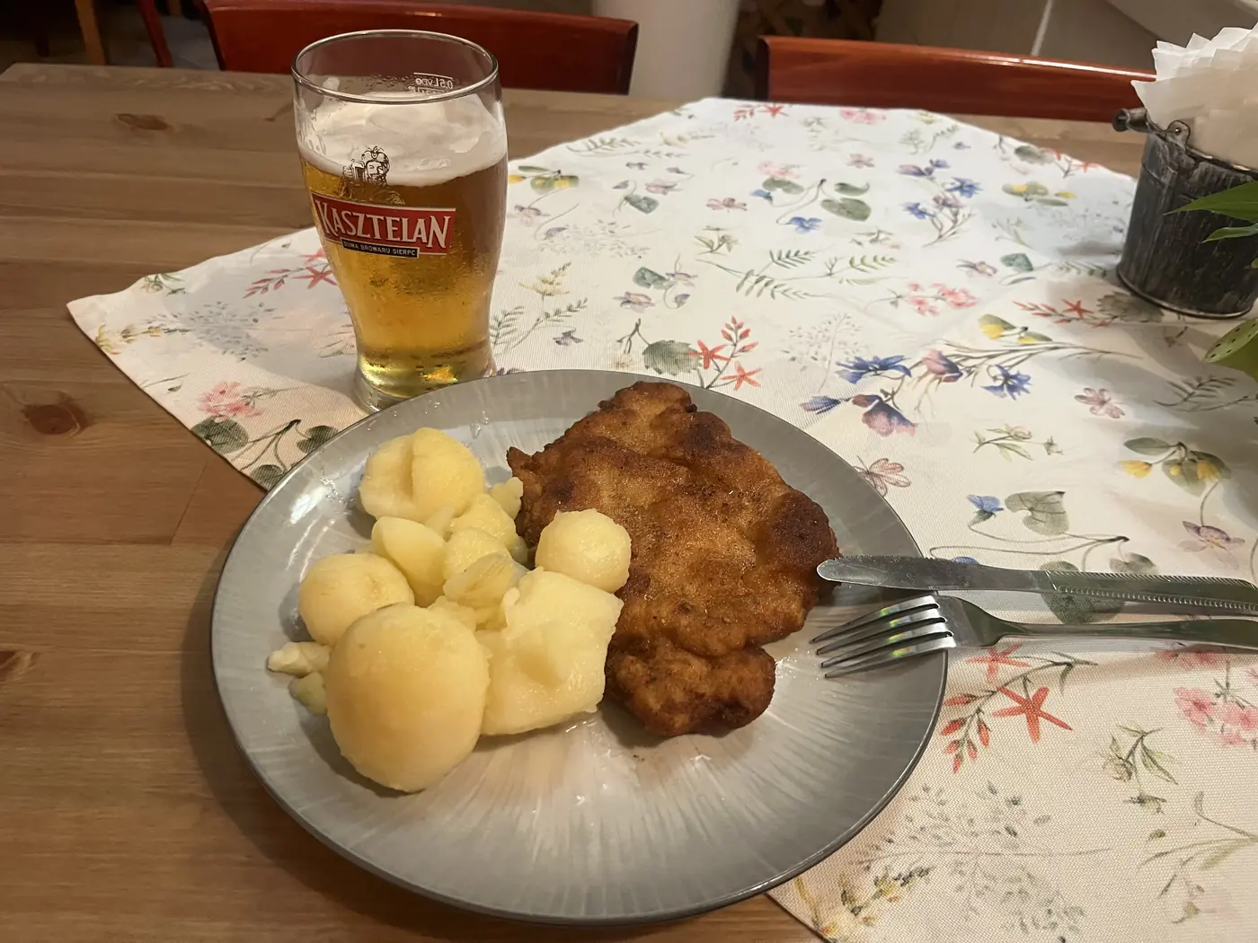 Polish dinner of breaded pork schnitzel with boiled potatoes and a glass of Kasztelan beer on a floral tablecloth