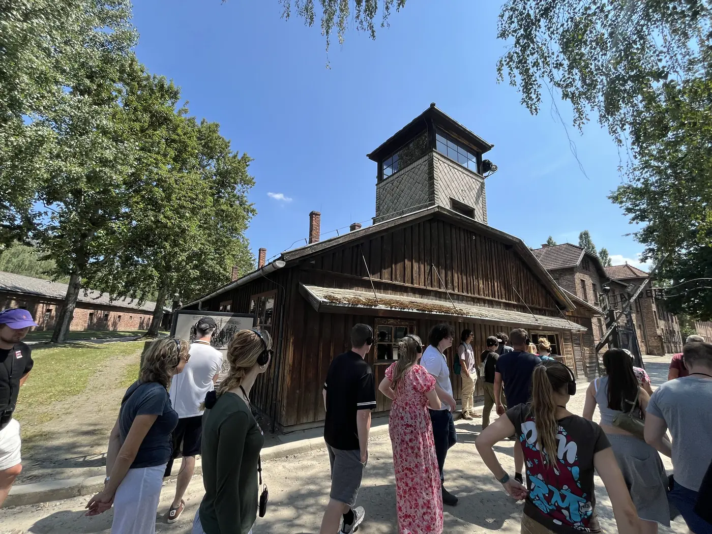 Visitors with audio guides walking past a wooden barrack building with a guard watchtower at the Auschwitz I memorial and museum