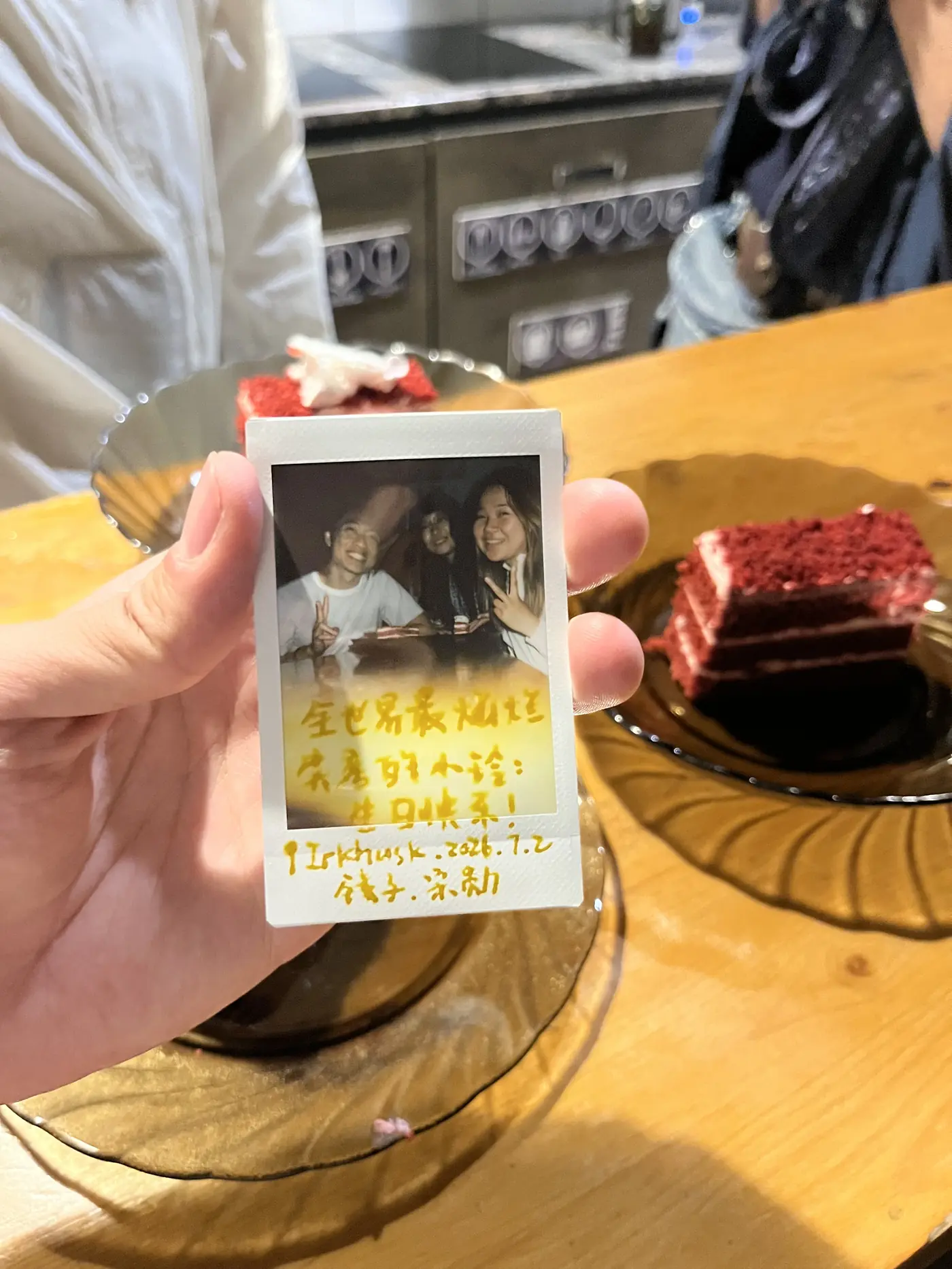 Hand holding a Polaroid photo of Shin and two friends with a handwritten Chinese birthday message, red velvet cake on table in Irkutsk