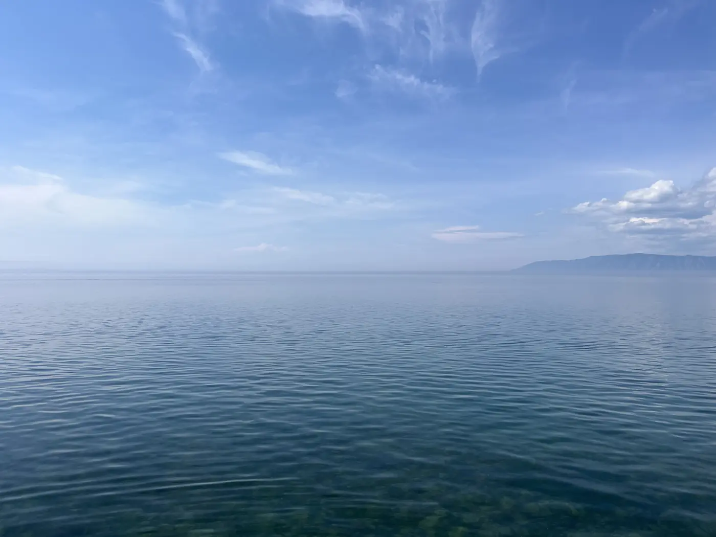Panoramic view of Lake Baikal from Listvyanka shoreline, deep blue water fading to hazy horizon with distant mountain silhouette, Siberia