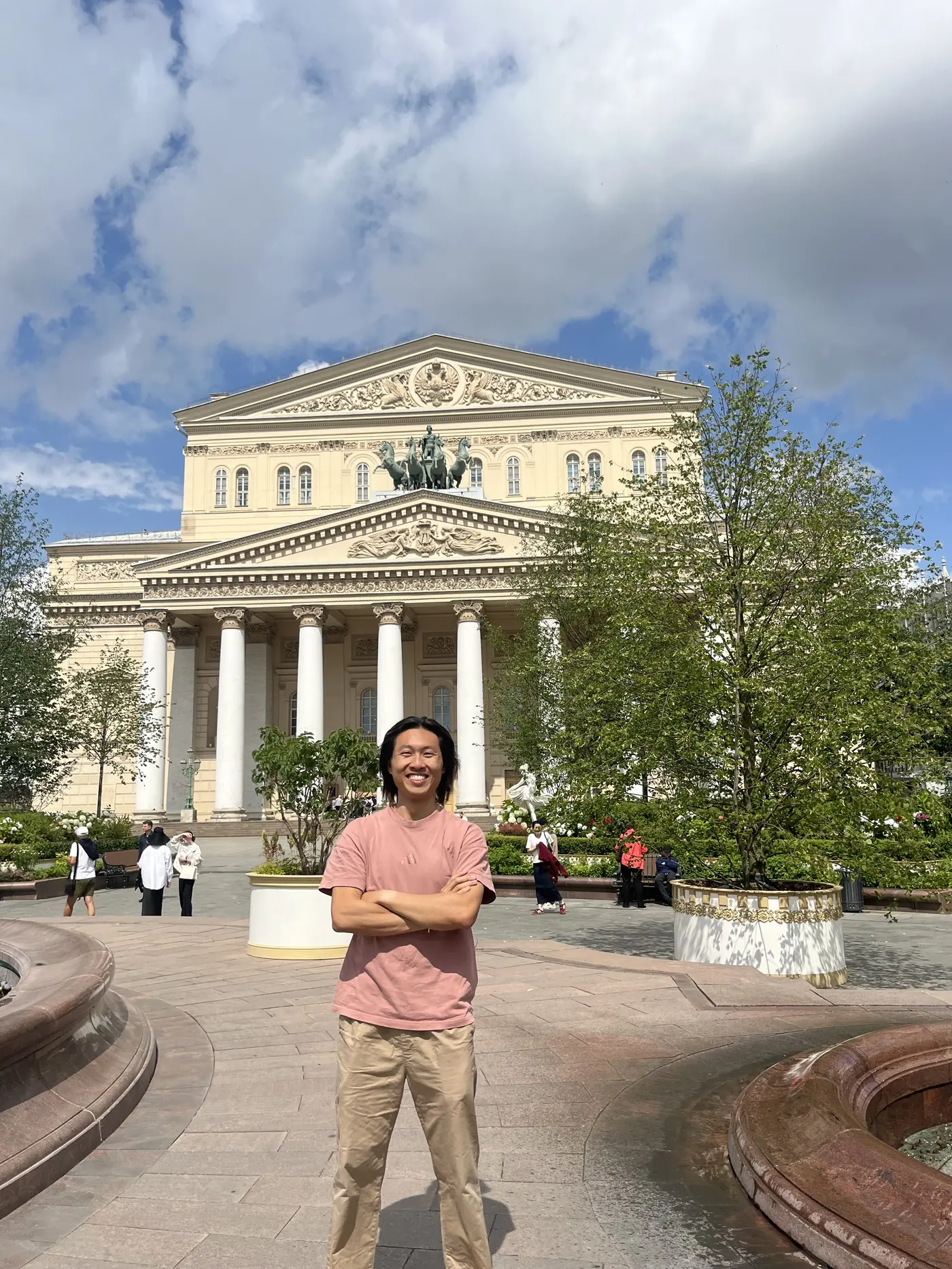Shin standing with arms crossed in front of the Bolshoi Theatre neoclassical facade, Moscow, summer day with clouds