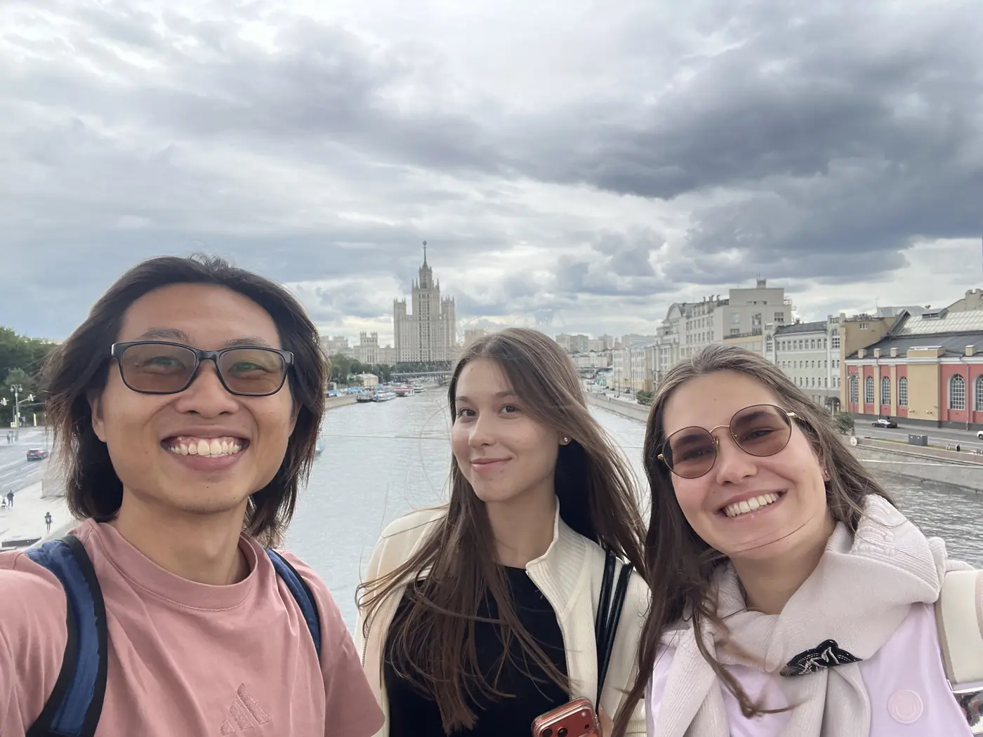 Shin selfie with two Russian women on a Moscow River bridge, Kotelnicheskaya building tower visible in the background, cloudy sky