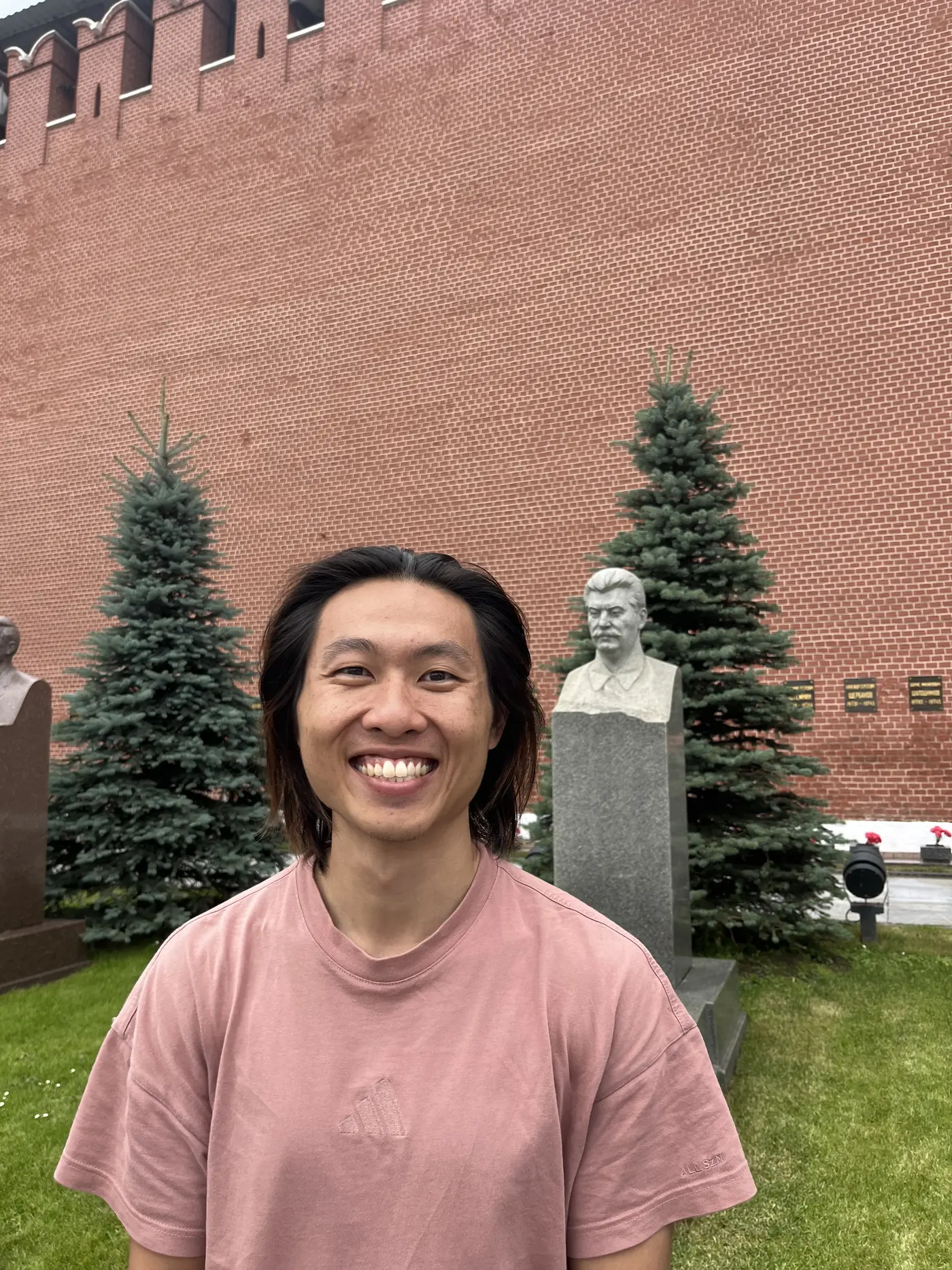 Shin smiling in front of the Kremlin wall necropolis, with a bust memorial and pine trees along the red brick wall, Moscow