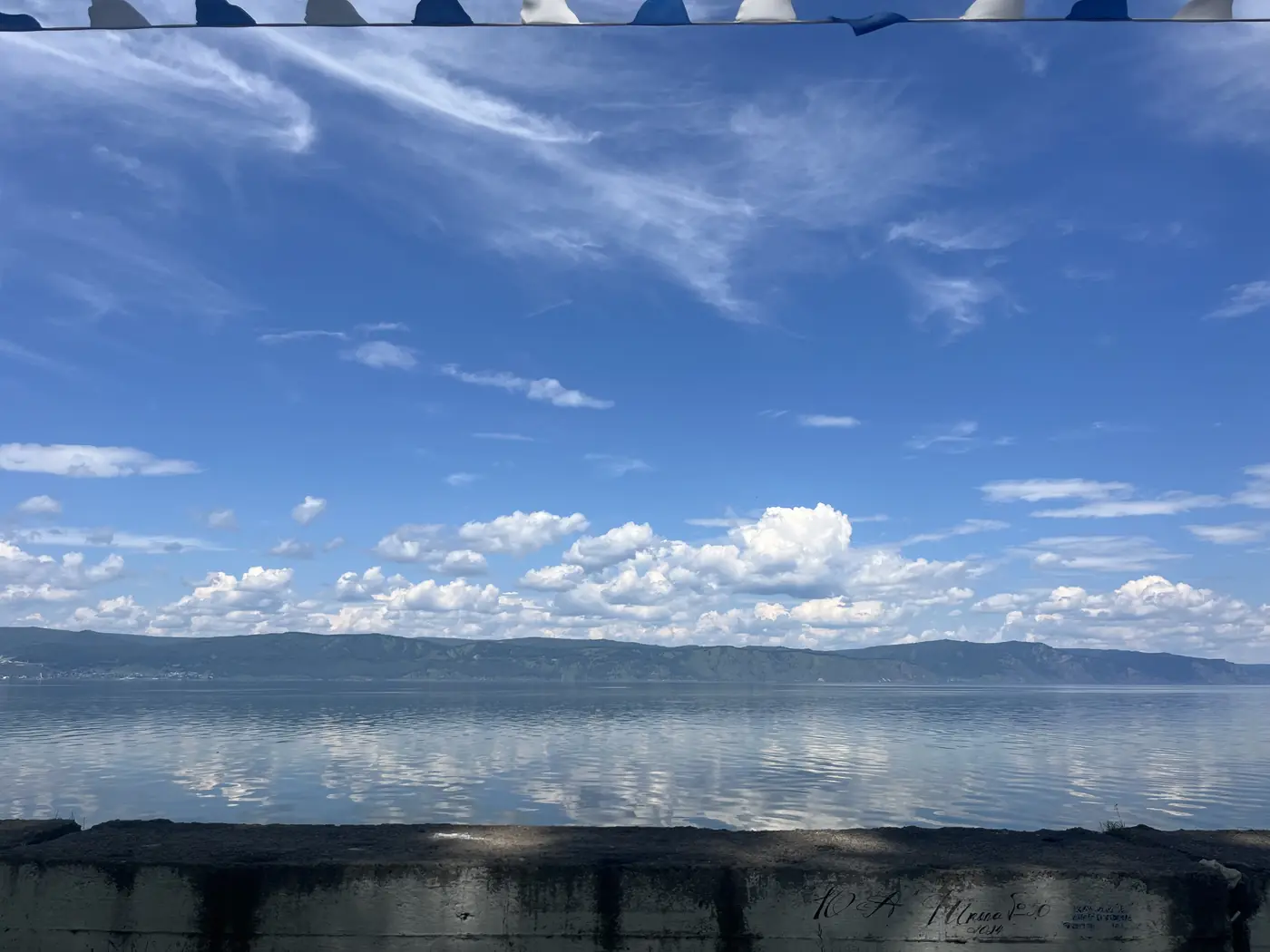 Lake Baikal view from Slyudyanka waterfront with bunting flags, calm water reflecting clouds and distant mountains, Siberia Russia