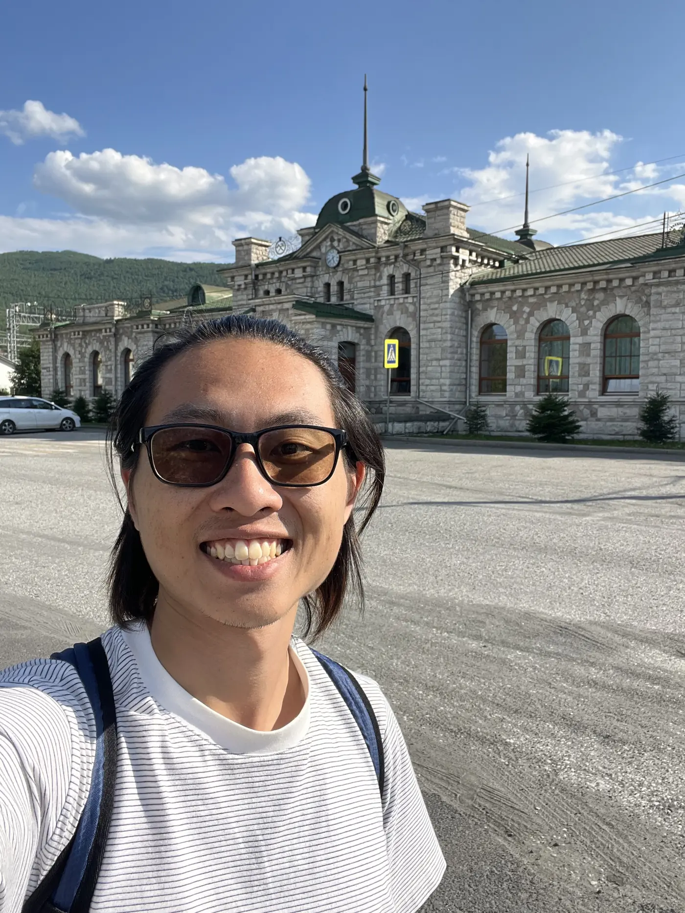 Shin selfie in front of Slyudyanka's historic marble railway station with arched windows and green dome, mountains behind, Lake Baikal Russia